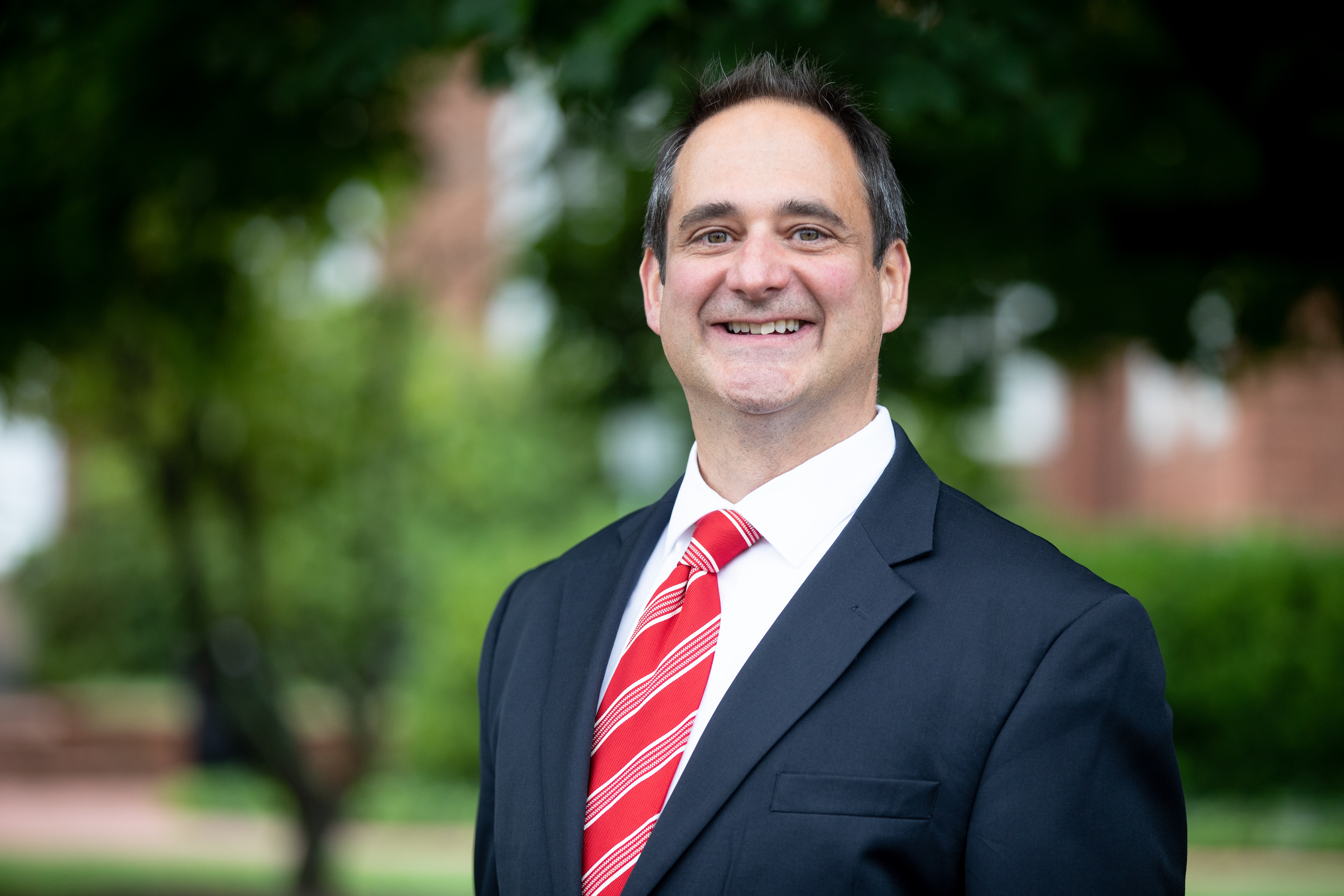 Dr. Vince Magnini - Portrait of a man in a dark suit and white shirt with a red striped tie. He smiles while standing outdoors in front of green trees. The background is softly blurred with hints of brick buildings. The image appears to be a professional headshot.