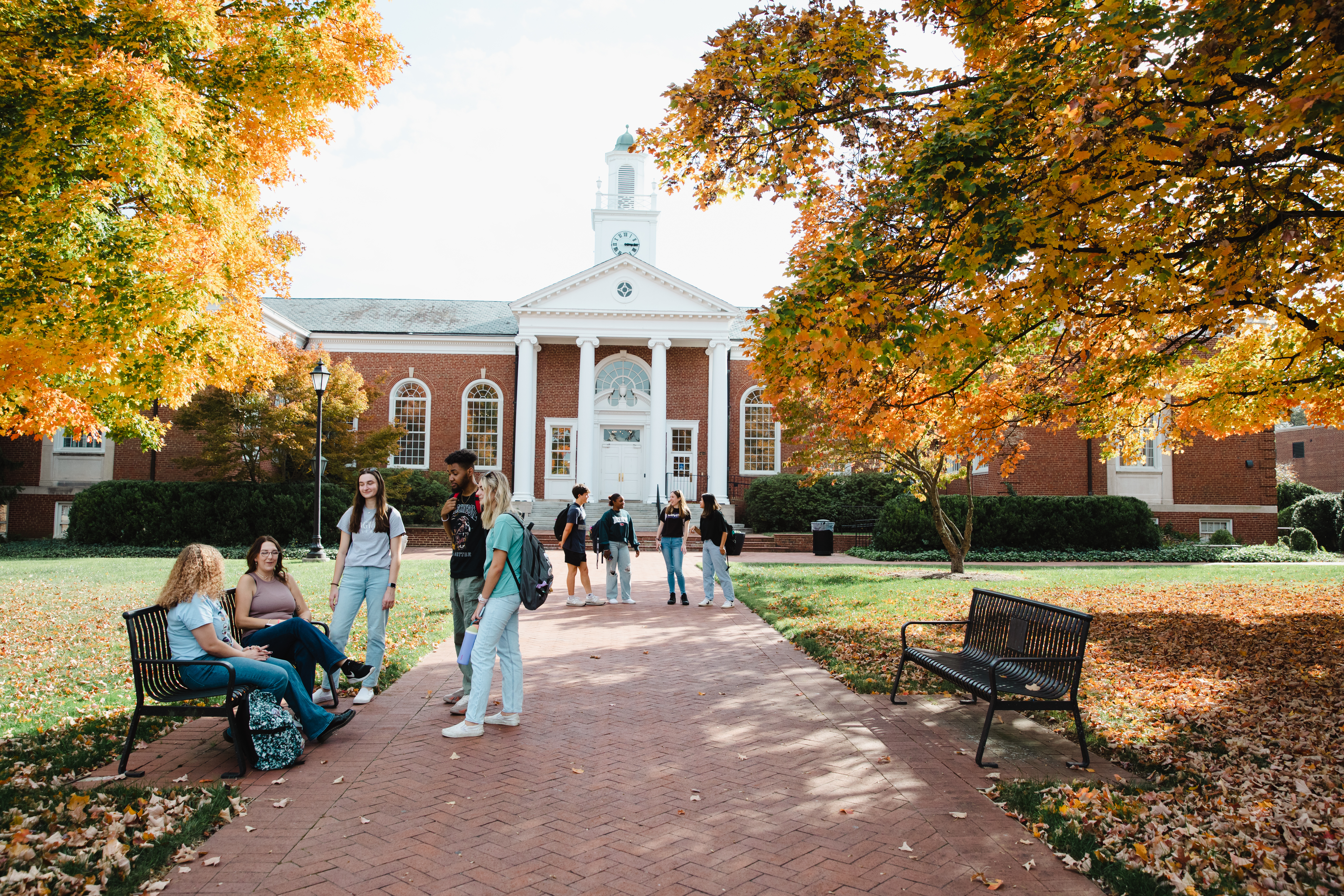 Students sitting and walking outside Eason Hall in the fall - Students gather and walk along a brick path in front of a red-brick academic building with white columns and a clock tower. Some sit on benches while others stand and talk between classes. Golden and orange fall leaves frame the scene and cover the ground. The campus setting reflects a bright autumn day.