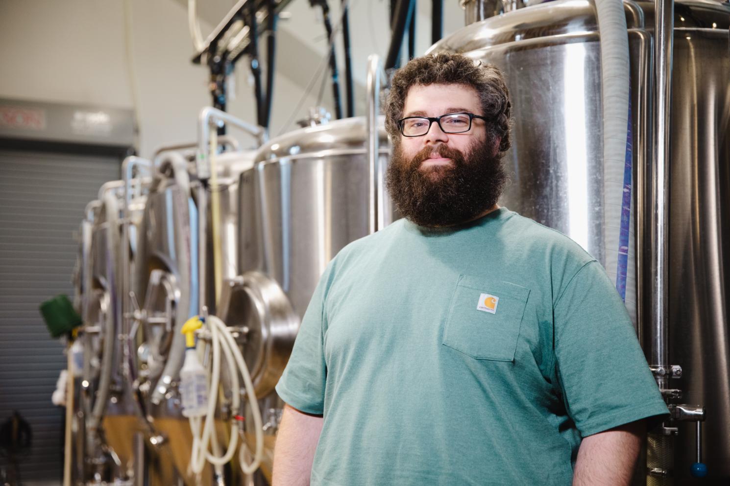 Brian Mandeville '10 - Man with glasses and a full beard stands inside a brewery next to large stainless steel tanks. He wears a green T-shirt and faces the camera with a slight smile. Metal pipes and hoses connect the brewing equipment behind him. The industrial space is softly lit with a roll-up door visible in the background.