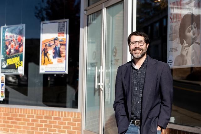 Eddie Mullins - Man wearing glasses, a dark blazer, and jeans stands outside the High Street Theatre entrance. Movie posters are displayed in the windows beside the glass doors. Brickwork lines the base of the building. Bright sunlight casts strong shadows across the storefront.