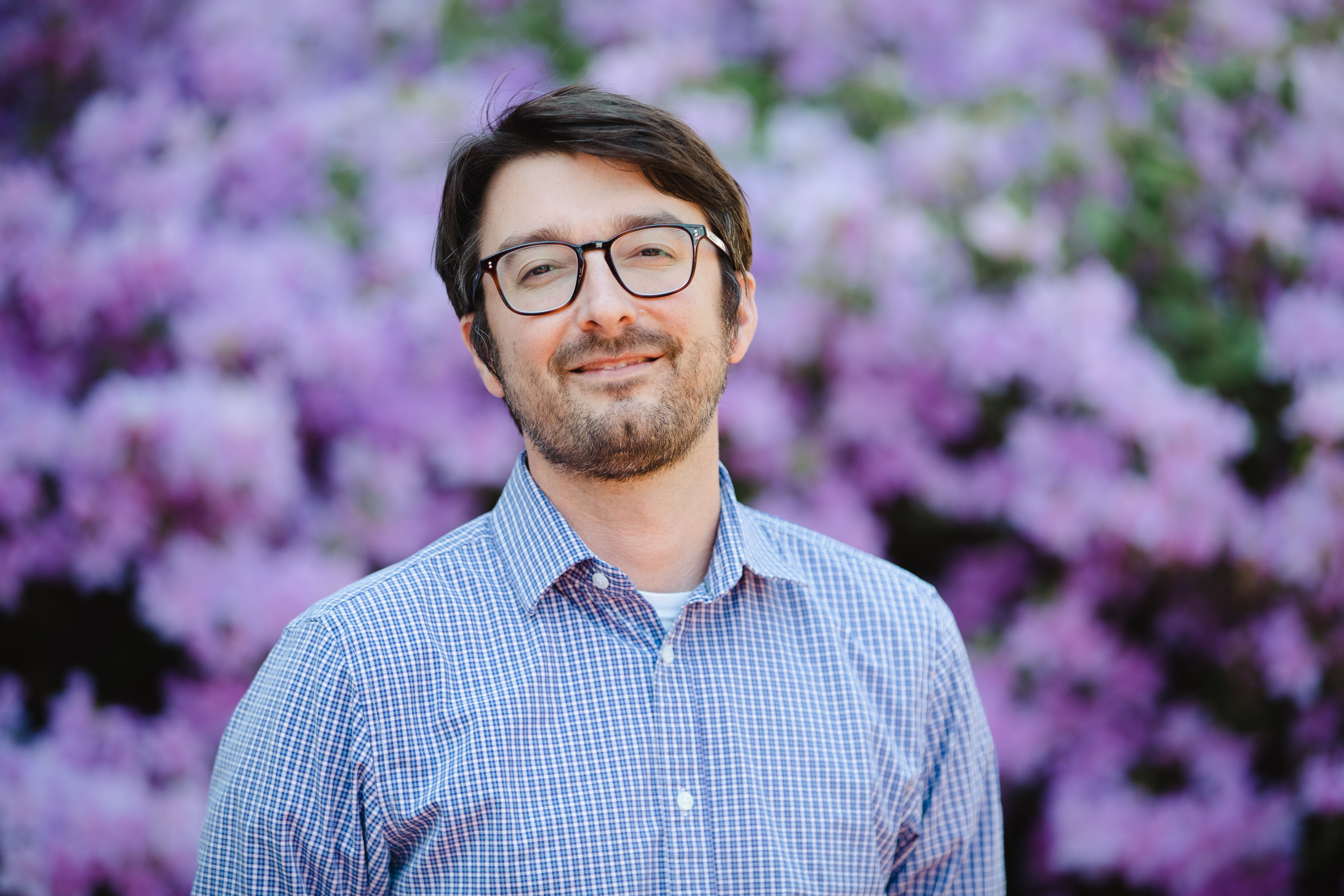 Alex Grabiec ’07 - Man with glasses stands in front of blooming purple flowers. He wears a blue checkered button-down shirt. He smiles gently at the camera. The background is softly blurred with vibrant blossoms.