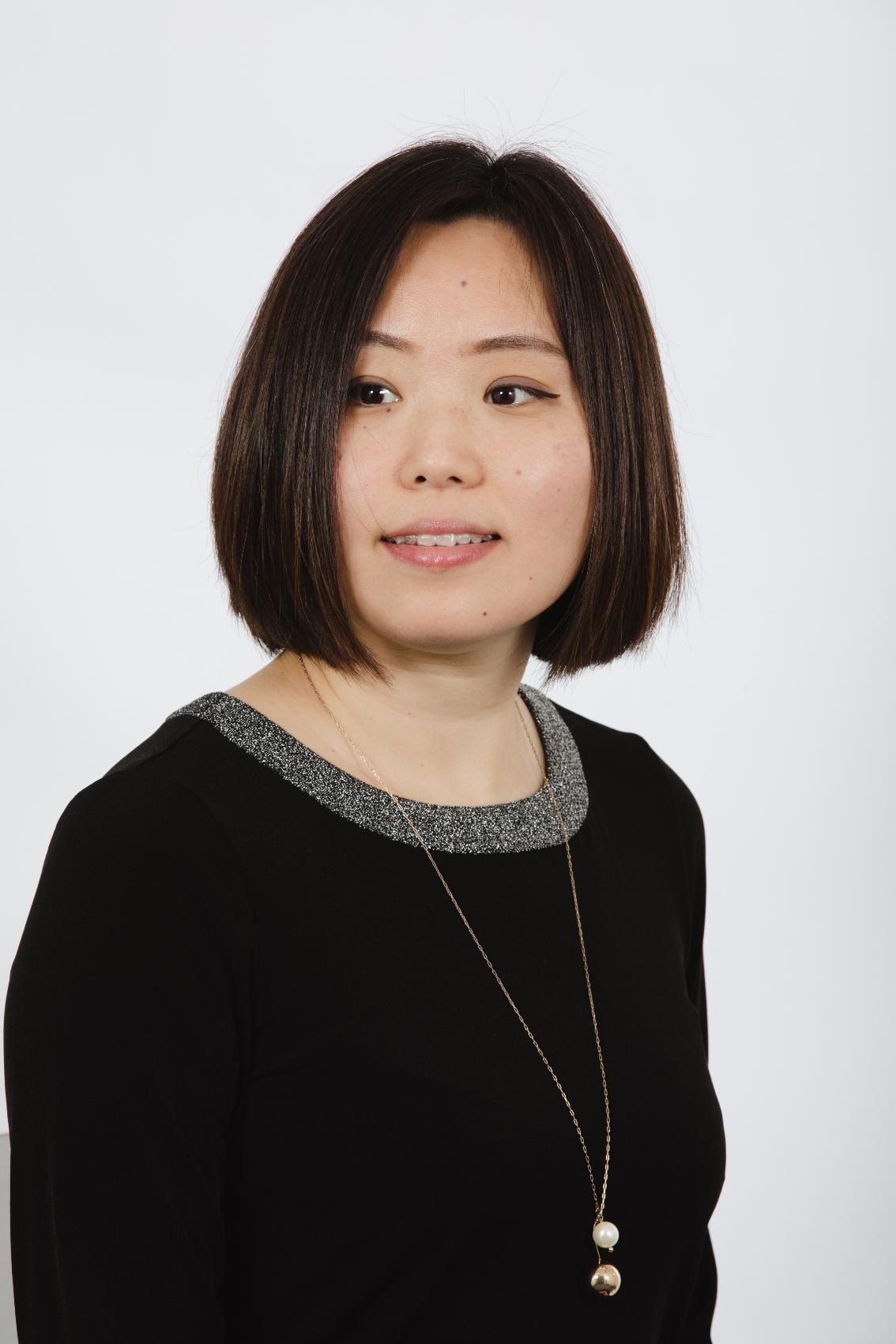 Dr. Meg Michelsen - Woman poses in a studio portrait against a light gray background. She wears a black top with a silver-trimmed neckline and a long pendant necklace. Her short dark hair frames her face as she looks slightly to the side. Professional head-and-shoulders image with a calm expression.