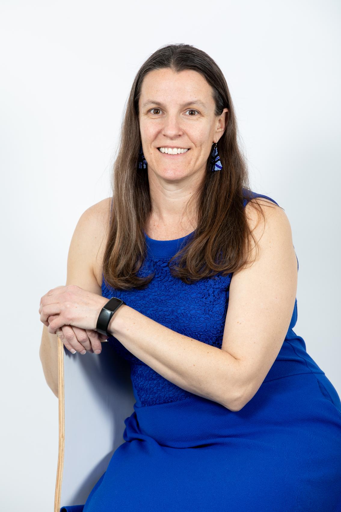 Dr. Melissa Kravetz - Woman in a blue sleeveless dress poses against a light gray background. She rests her arms on the back of a wooden chair and smiles at the camera. She wears blue earrings and a black smartwatch. Professional studio portrait with a friendly, confident expression.