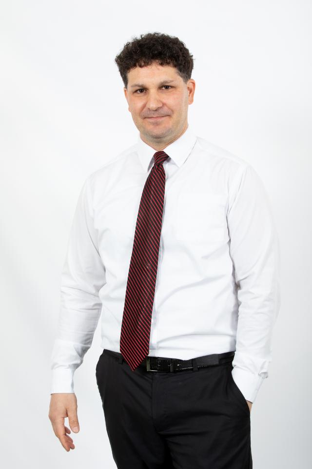 Dr. Mike Mucedola, associate professor of health and physical education - Man stands against a light gray studio background. He wears a white dress shirt, black pants, and a red striped tie. One hand rests in his pocket as he looks toward the camera. Professional portrait with a neutral expression and relaxed stance.