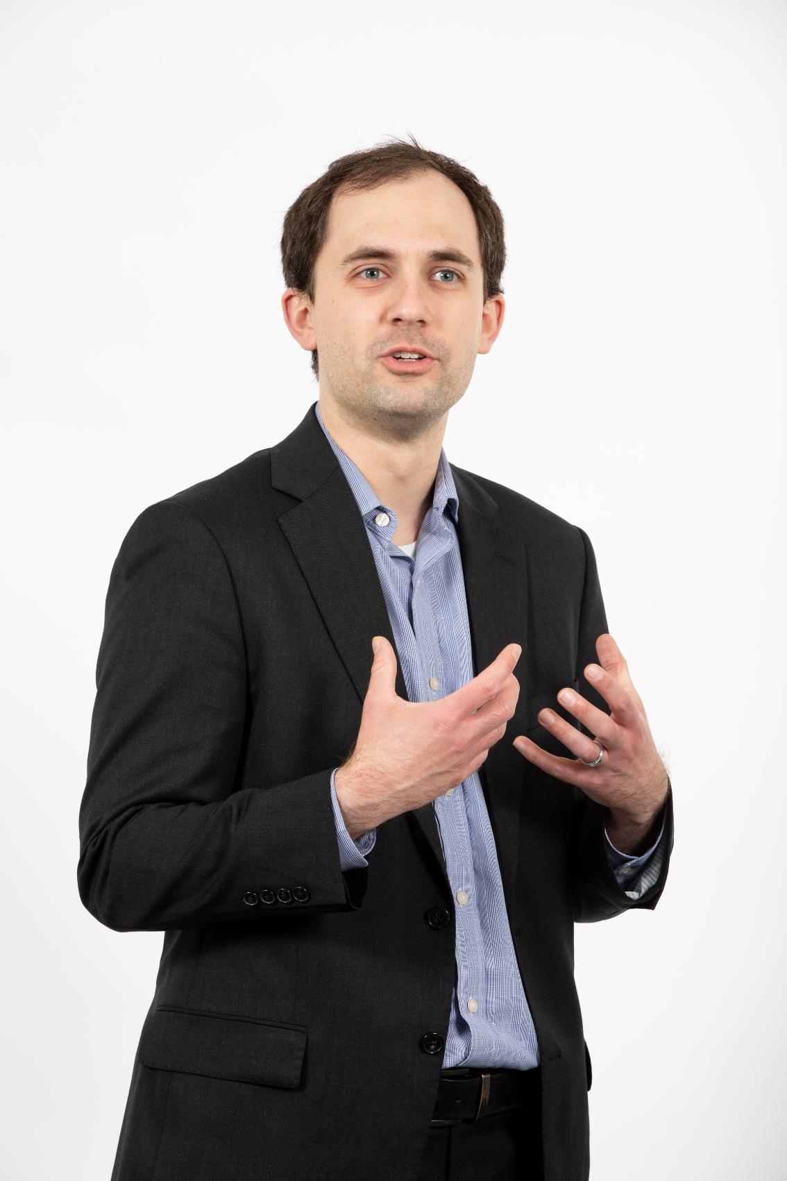 Dr. Mike Waddell, assistant professor of music - Man in a dark blazer and light blue shirt poses against a light gray background. He gestures with both hands as if explaining something. He looks slightly off camera with a focused expression. Professional studio portrait capturing an engaged speaking moment.
