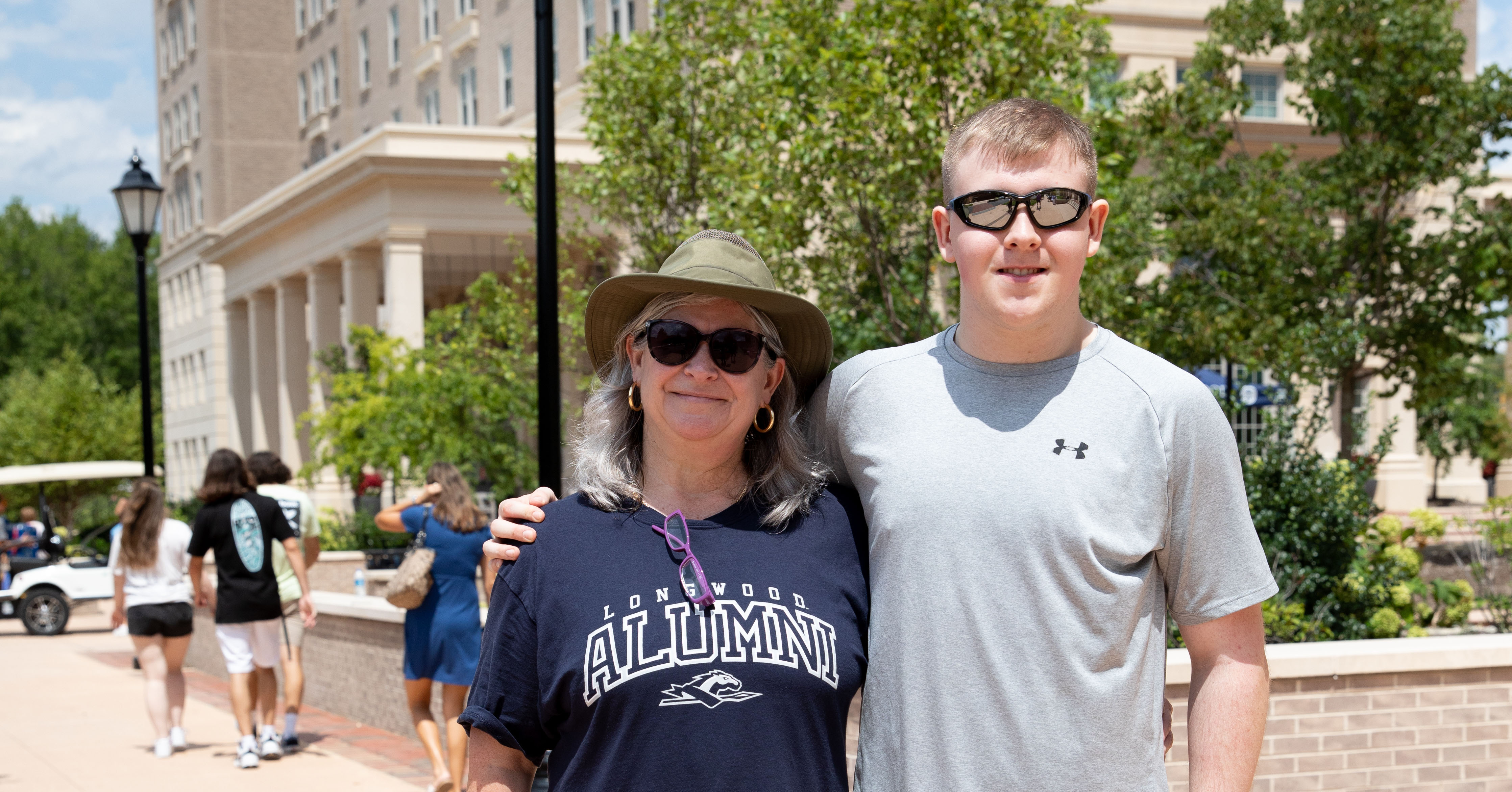 Grey Sweeney ’86 and William Perkins ’27 on move in day - Woman wearing a “Longwood Alumni” T-shirt stands with a young man on campus. Both wear sunglasses and smile with arms around each other. Campus buildings and trees are visible behind them. Other students walk along the sidewalk in the background.