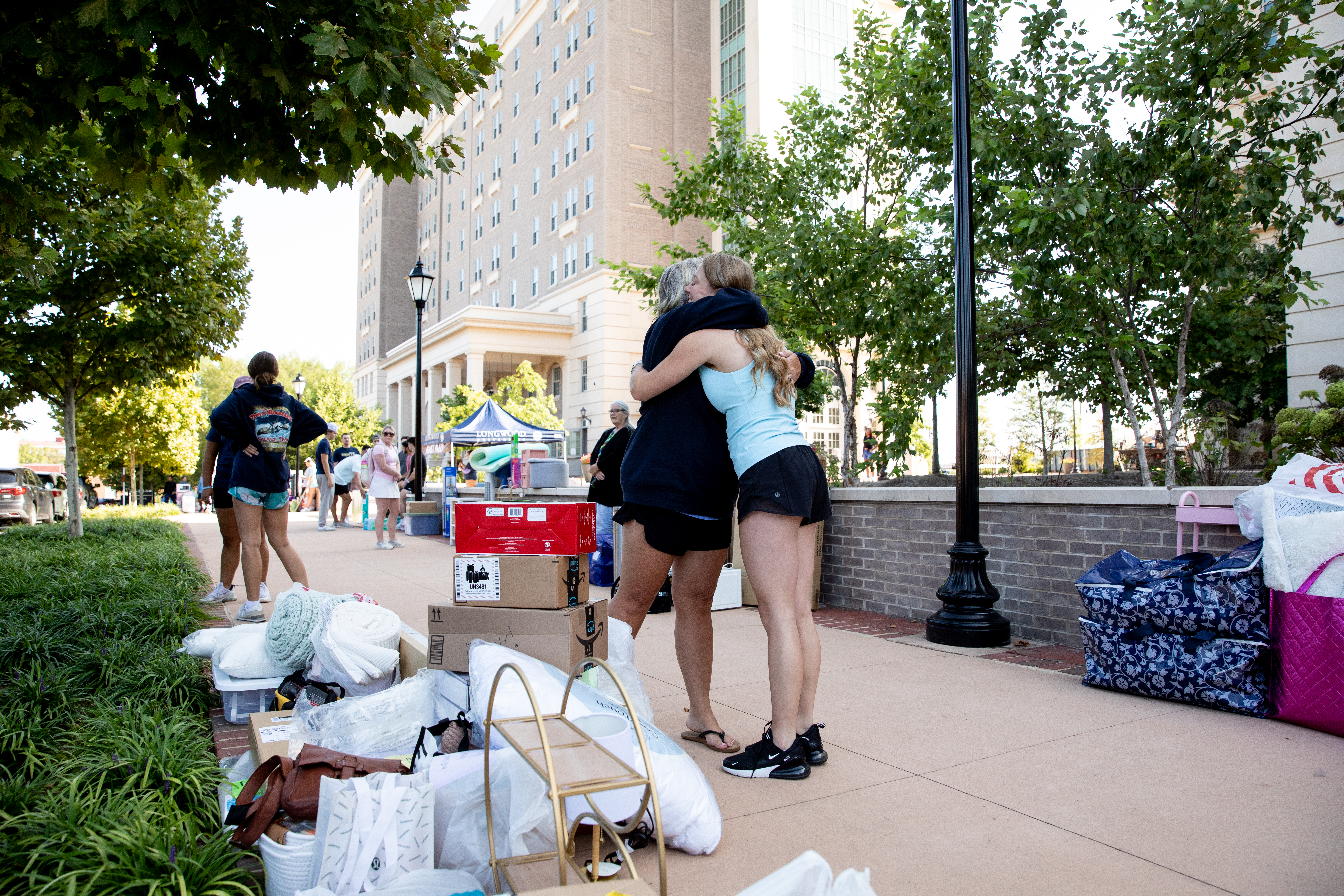 A hug outside halls during move-in 2024 - Two students hug on a sidewalk outside a campus residence hall during move-in day. Boxes, bedding, and bags are stacked nearby. Other students and families gather along the walkway. Trees and a tall brick residence hall rise in the background.