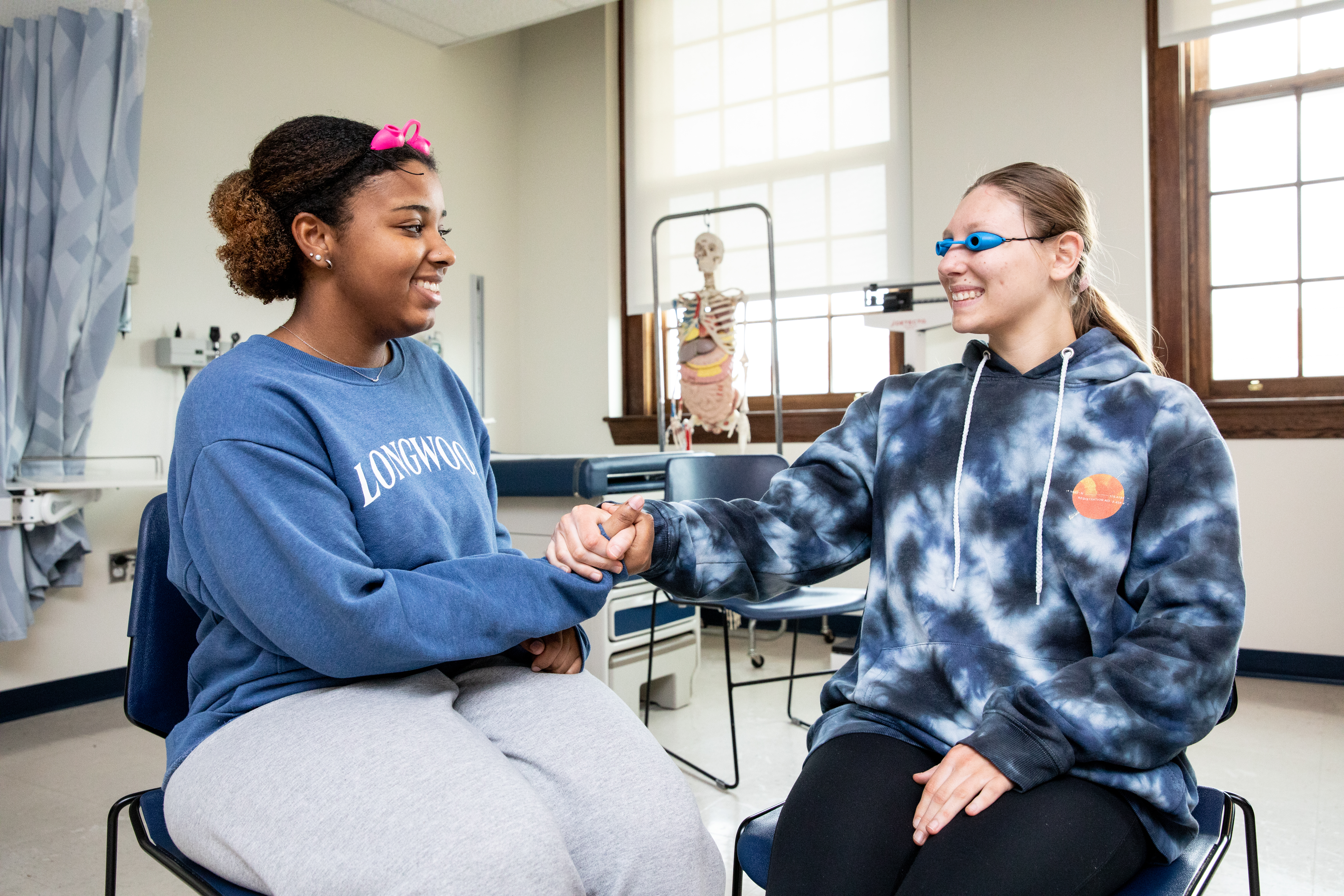 Nursing students participating in Experiential Dementia Skills Training - Two students sit facing each other in a clinical skills classroom. One student wearing blue sensory goggles shakes hands with the other. A skeleton model and medical equipment are visible behind them. They smile while practicing a hands-on healthcare exercise.