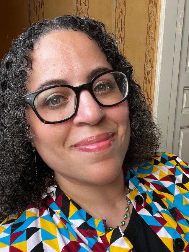 Kiki Petrosino - Close-up portrait of a woman with short curly hair and black-framed glasses. She smiles softly while wearing a colorful geometric-patterned jacket. A small nose stud and necklace are visible. The background shows a warm-toned wall and a doorframe.