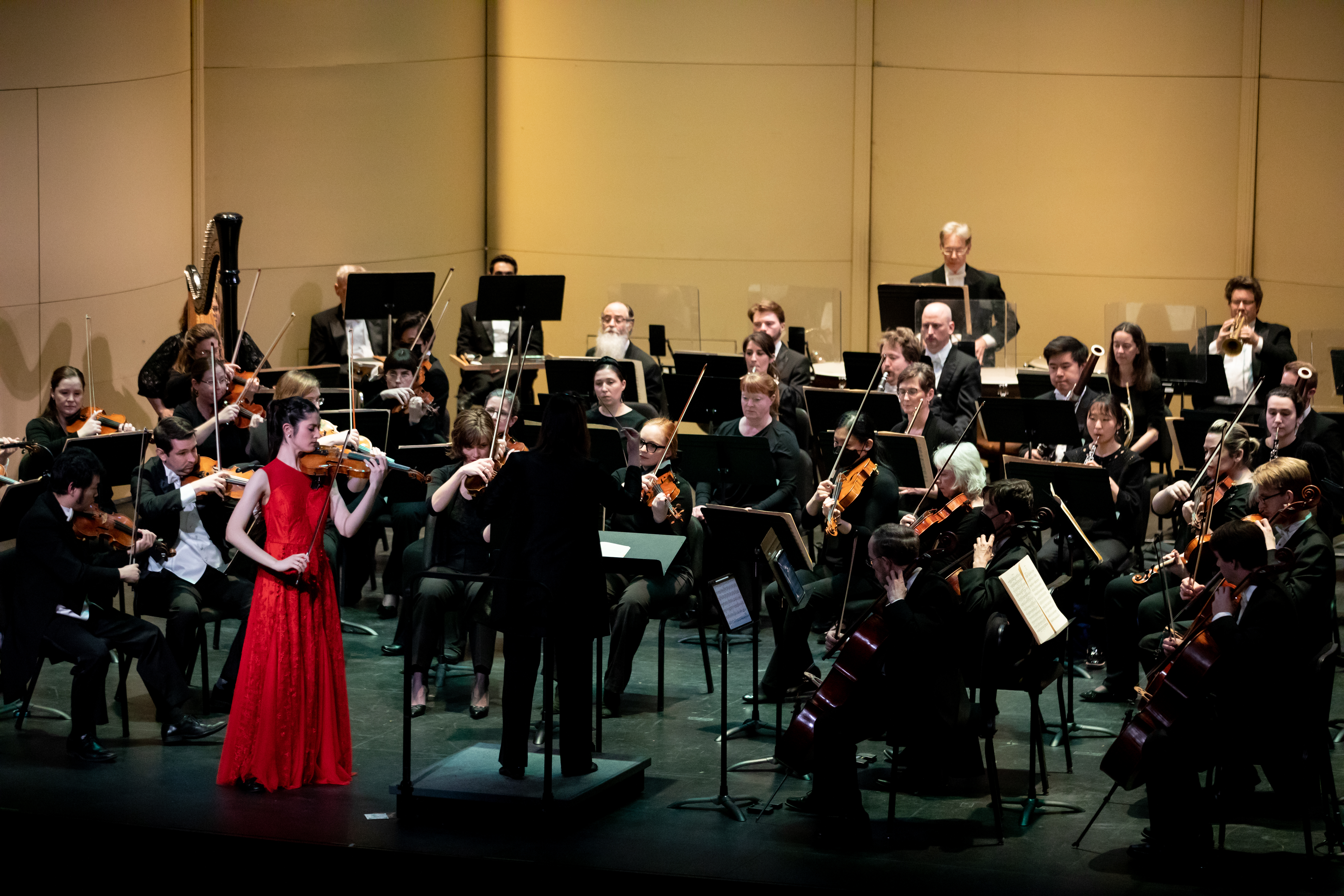 Richmond Symphony Orchestra performing on campus earlier in 2024 - Orchestra performs on stage with a conductor leading from the podium. A violin soloist in a bright red gown stands at the front of the ensemble. Musicians in black concert attire play string and brass instruments. The performance takes place in a formal concert hall.