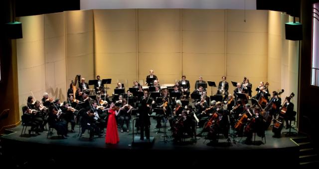 Richmond Symphony at Longwood University - Full orchestra performs on a concert hall stage. A conductor leads the musicians from the podium. A solo violinist in a bright red gown stands at the front. String, brass, woodwind, and percussion sections fill the stage.