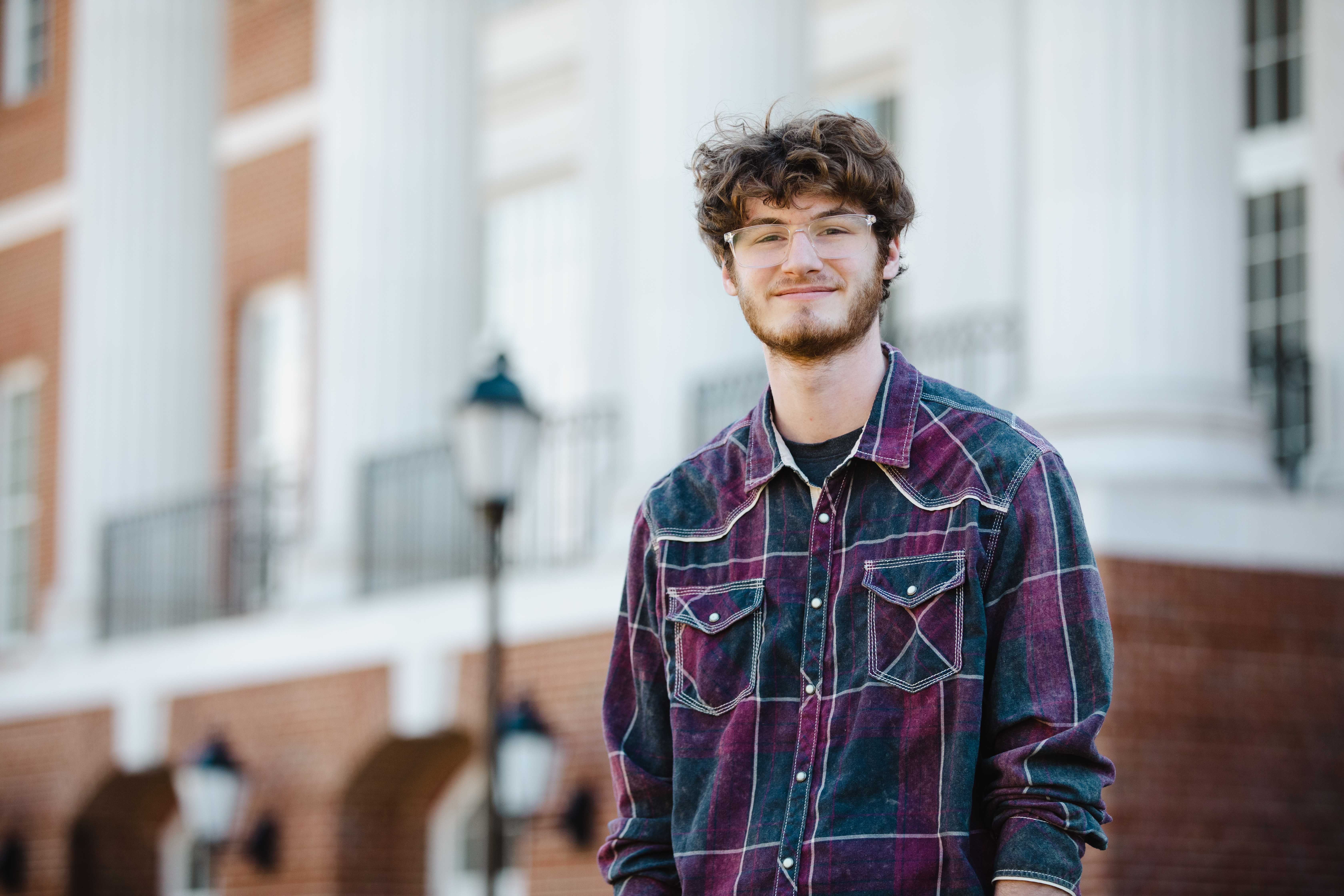 Ryan Urban ’26 - Young man wearing glasses and a plaid button-down shirt stands outdoors on campus. He smiles slightly while facing the camera. White columns and red-brick walls of a campus building rise behind him. A black lamppost and iron railing are visible along the steps in the background.