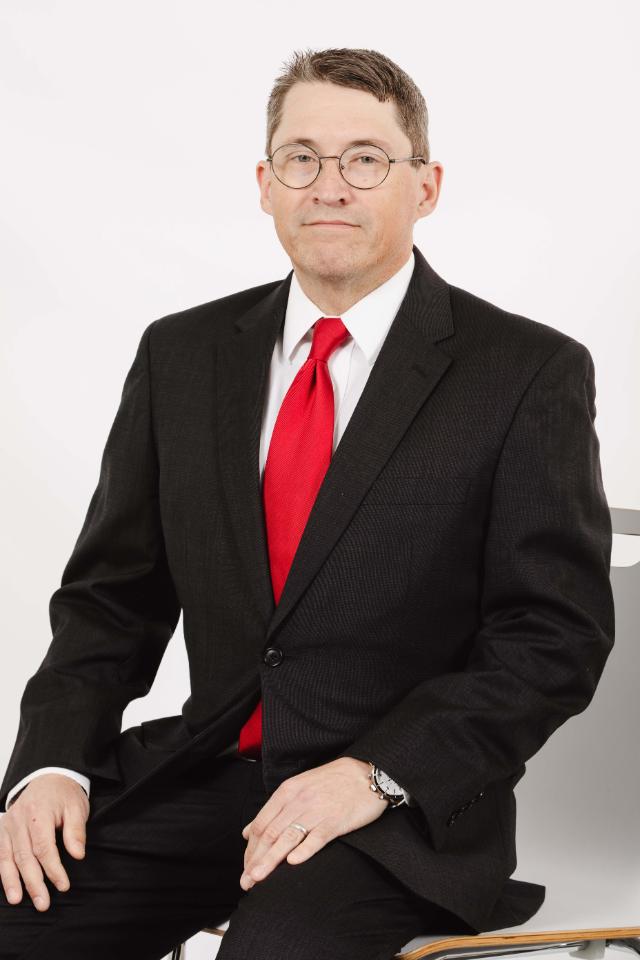 Dr. Shawn Smith - Man in a dark suit and red tie sits against a light gray background. He wears glasses and rests his hands on his knee. He looks directly at the camera with a neutral expression. Professional studio portrait with a formal appearance.