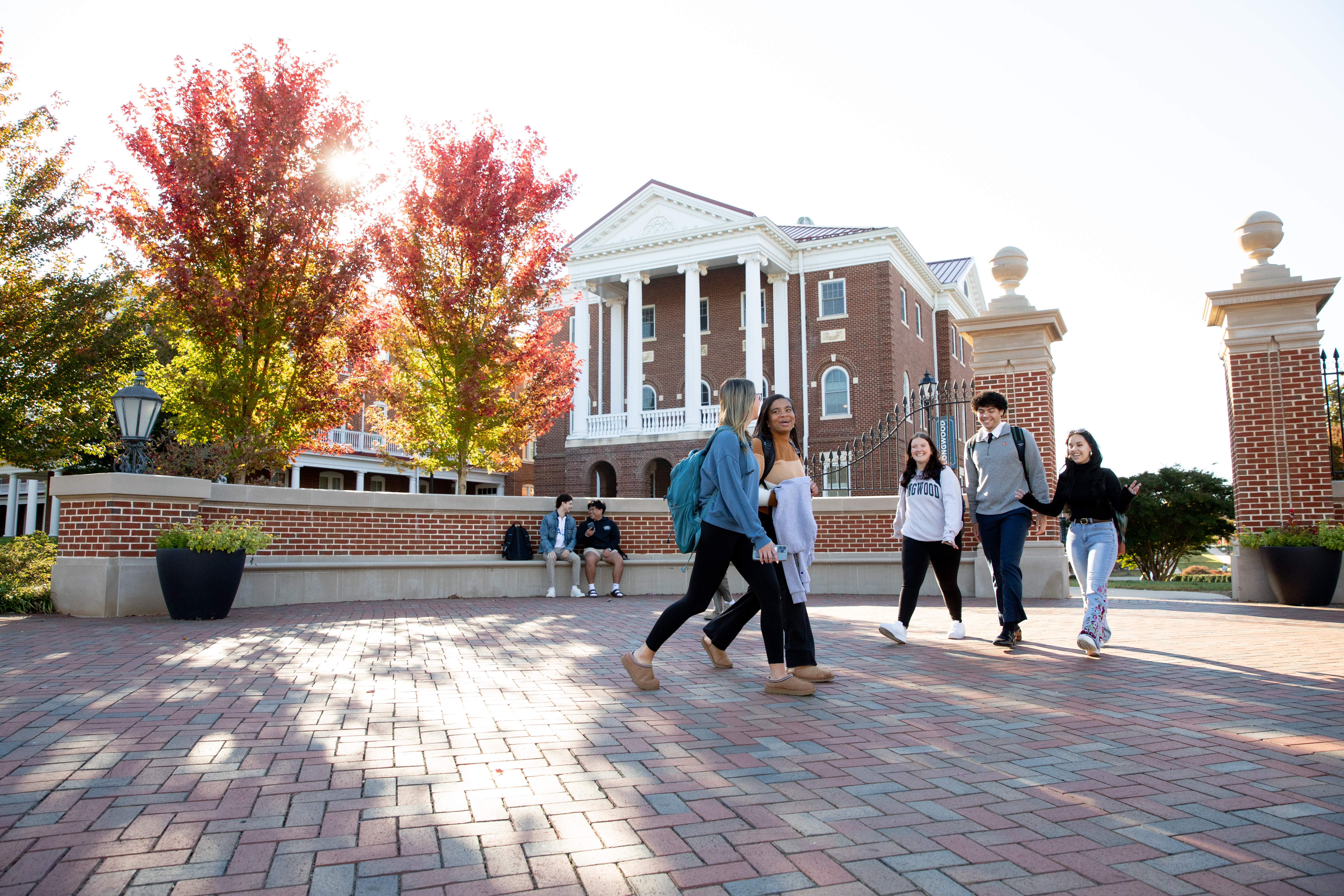 Students walking in front of The Gate at Longwood on the way to class - Students walk across a brick plaza in front of a red brick academic building with white columns. Colorful fall trees frame the scene as sunlight shines through the leaves. Two students sit on a low brick wall in the background. The group appears engaged in conversation while heading across campus.