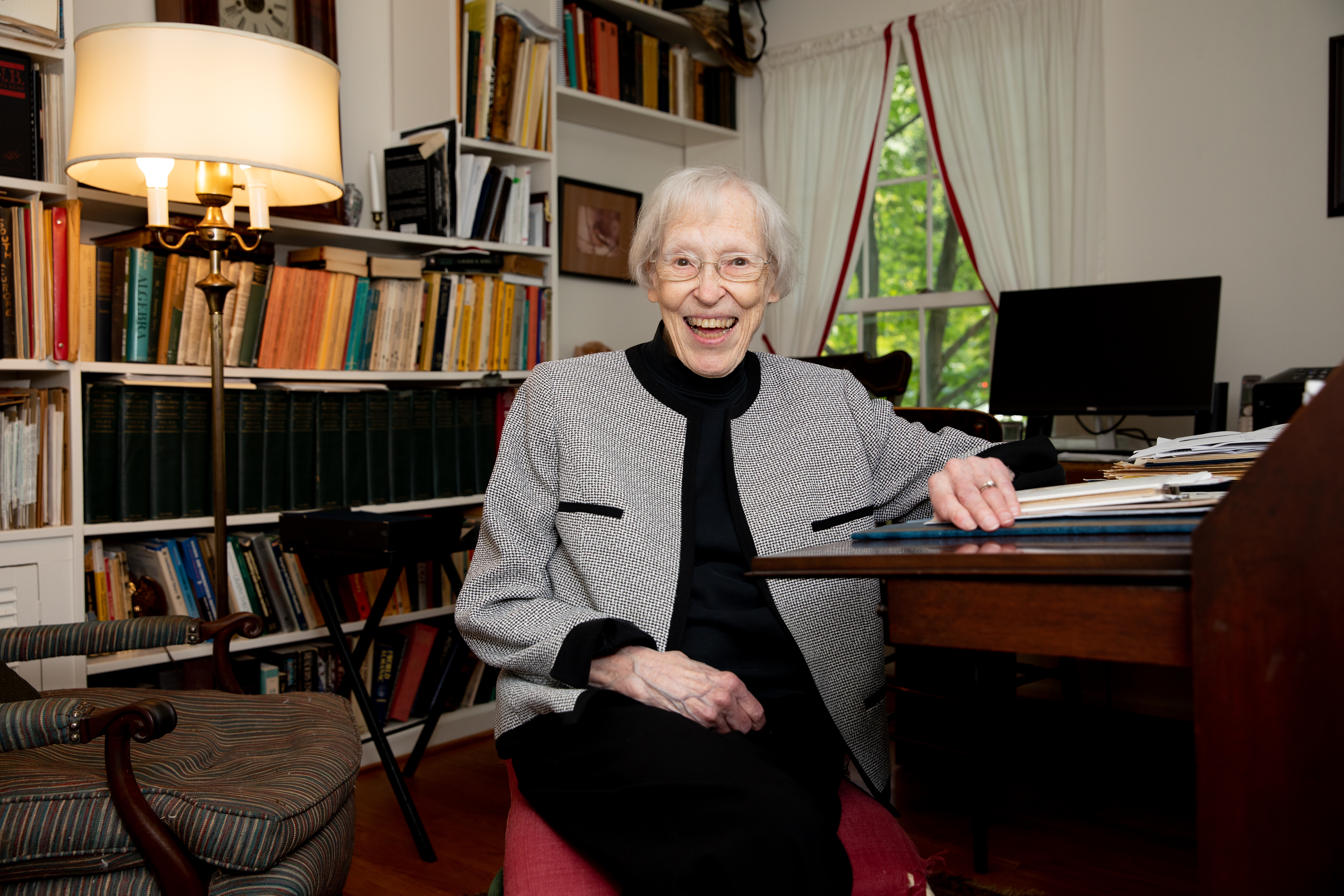 Dr. Susan May - Elderly woman smiles while seated at a wooden desk in a home office. Bookshelves filled with books line the wall behind her. A lit table lamp and framed artwork add warm light to the room. A window with white curtains overlooks green trees outside.