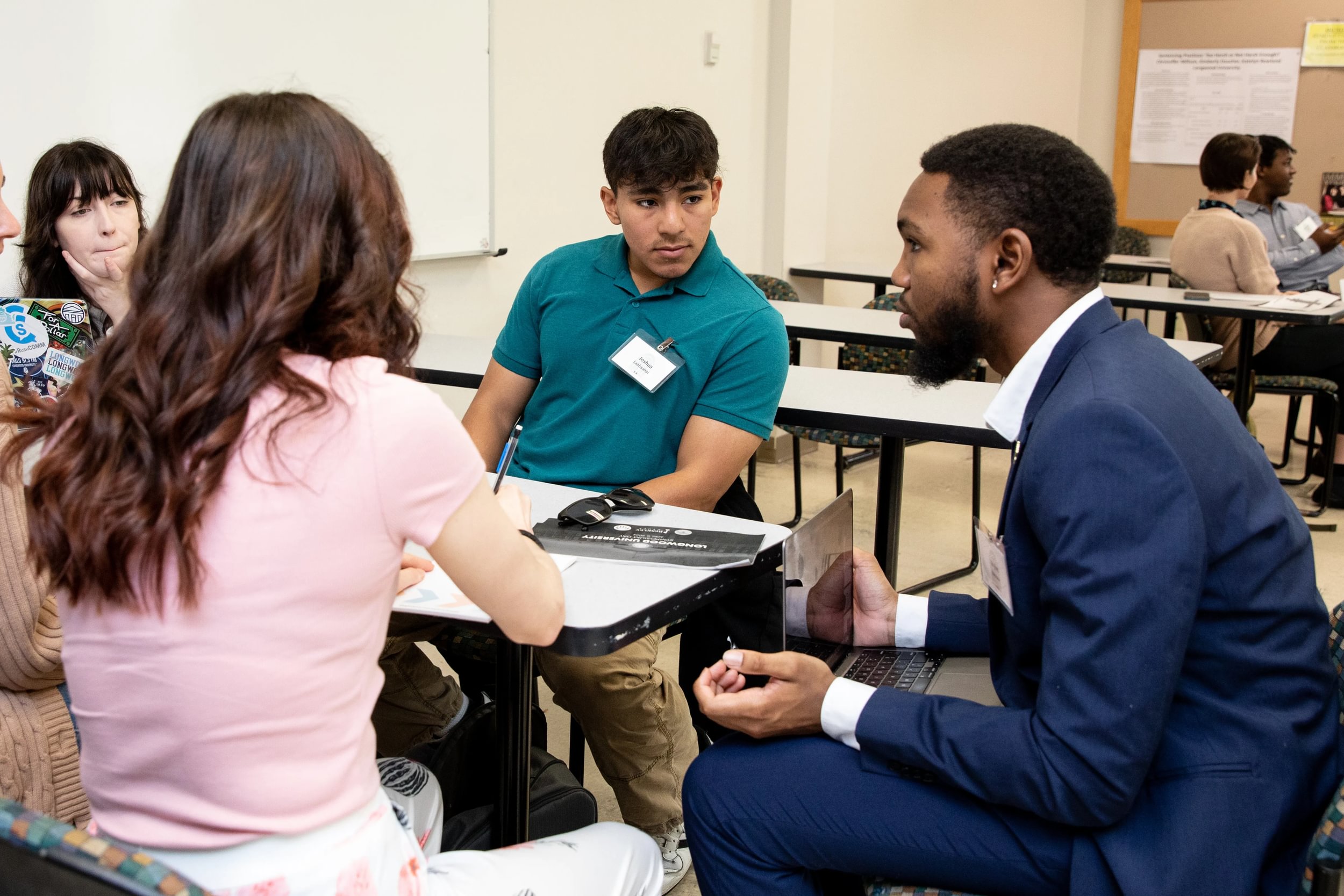 Students deliberate on citizenship and the issues citizens face in their communities on Symposium Day, the capstone of Longwood's Civitae Core Curriculum. - Four participants engage in small-group discussion during a campus symposium. A professional in a blue suit speaks while holding a laptop. Students with name tags listen and take notes at classroom desks. Other groups collaborate in the backgr