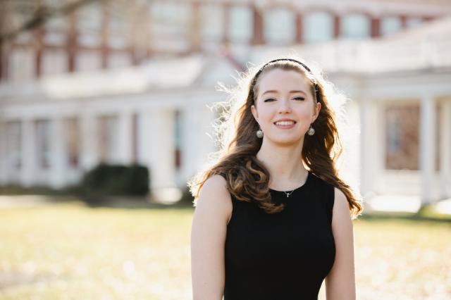 Tess Robertson ’25 - Young woman in a sleeveless black dress stands outdoors in front of a large campus building. Sunlight creates a soft glow around her hair as she smiles toward the camera. She wears drop earrings and a delicate necklace. The lawn and white columns of the building blur in the background.