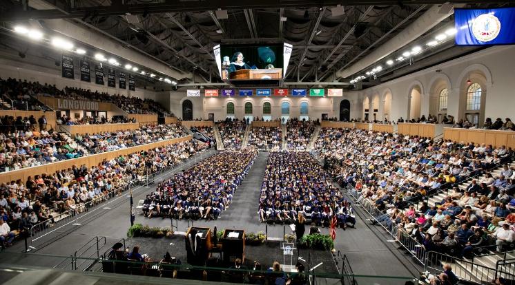 The undergraduate ceremony was held inside the Joan Perry Brock Center because of forecasted storms - A panoramic view of an undergraduate commencement ceremony in an arena. Graduates in blue caps and gowns fill the floor seating. Family and friends pack the surrounding stands. A large screen above the stage displays the speaker at the podium.