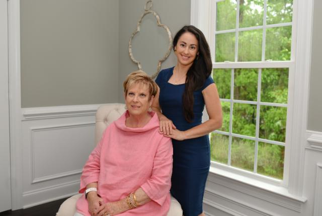 Rachel Nunnally Overstreet ’74, M.A. ’84 and Emily Wilson Overstreet ’06 - Two women pose for a portrait in a bright room by a window. One sits in a chair wearing a pink top while the other stands beside her in a blue dress. They smile toward the camera with greenery visible outside the window. Decorative wall molding and a framed mirror accent the space.