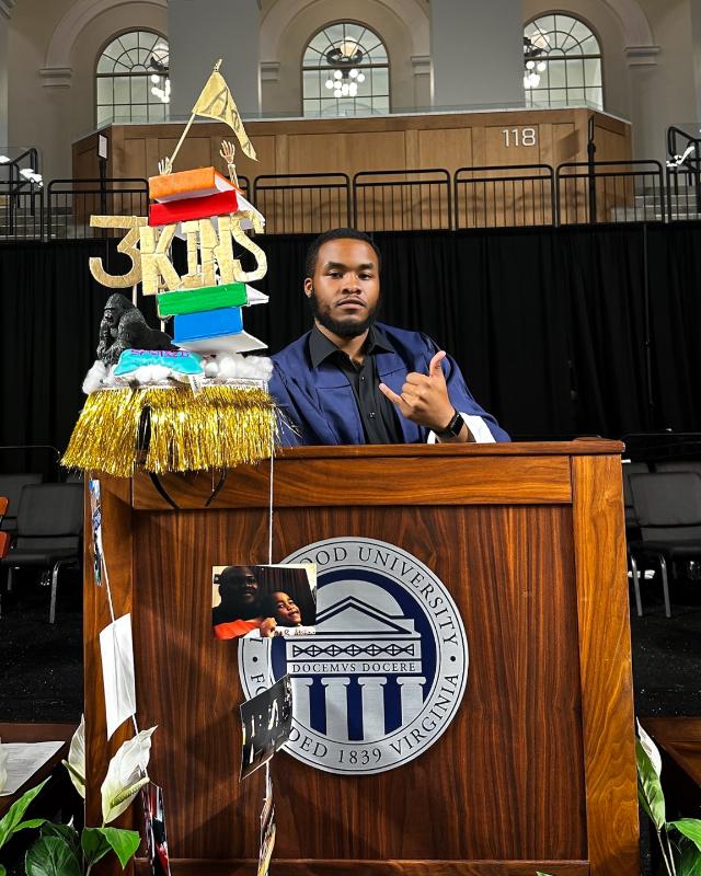 Jaylen Fedderman ’25 - Student stands behind a wooden podium with the Longwood University seal. A decorated graduation cap display with books, gold fringe, and “3KHS” sits beside him. He poses with a hand gesture on a stage inside a large arena. Arched windows and section number 118 appear in the background.