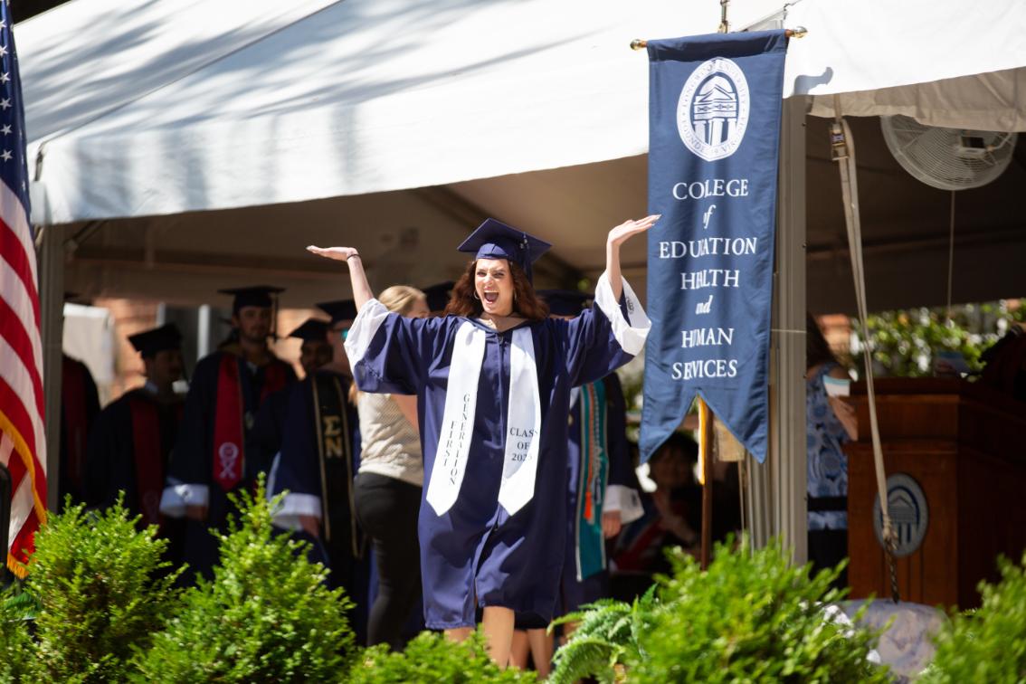 Graduate student wearing a first generation sash holds her hands in the air as she crosses the stage