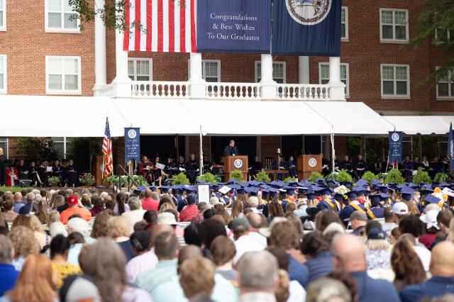 President W. Taylor Reveley, IV delivers his remarks - Crowd gathers for Longwood Commencement in front of a brick building. Graduates in blue caps and gowns face a stage under a large white tent. A speaker stands at the podium with faculty seated behind on the platform. American and Virginia flags hang above a banner congratulating graduates.