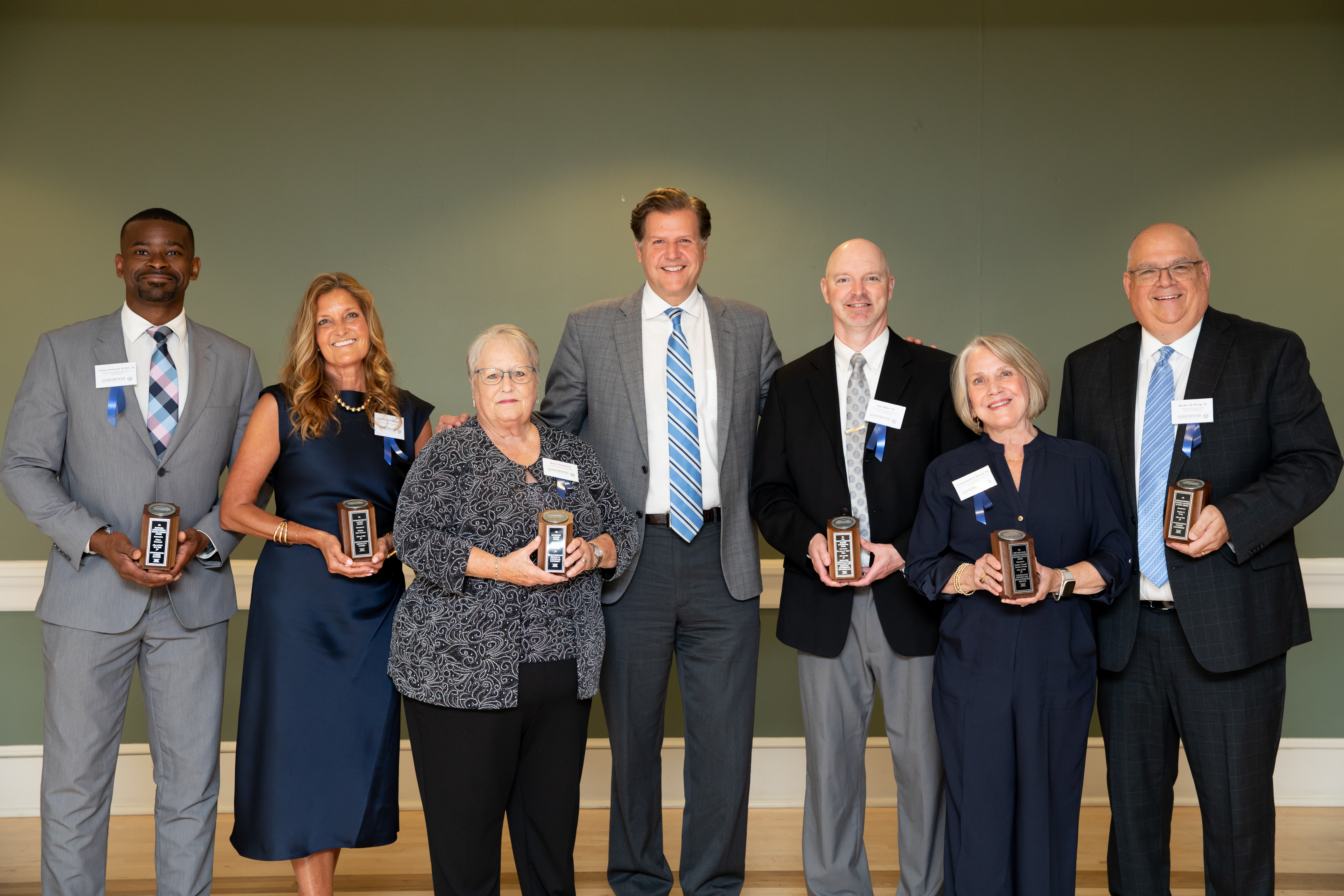 Pictured Left to Right: Cainan Townsend ’15, M.S. ’20; Michele Sims-Gannon ’90; Karen Mitchell Schinabeck, Hon. ’25; President W. Taylor Reveley IV; Dr. Jason “Jake” Milne ’99; Candice “Candy” Jamison Dowdy ’69; and Bradley “Brad” Pomp ’92. Not pictured: Elizabeth Stanton Kostelny ’81. - Seven people pose in a line indoors, several holding small award plaques. They wear business attire and name ba