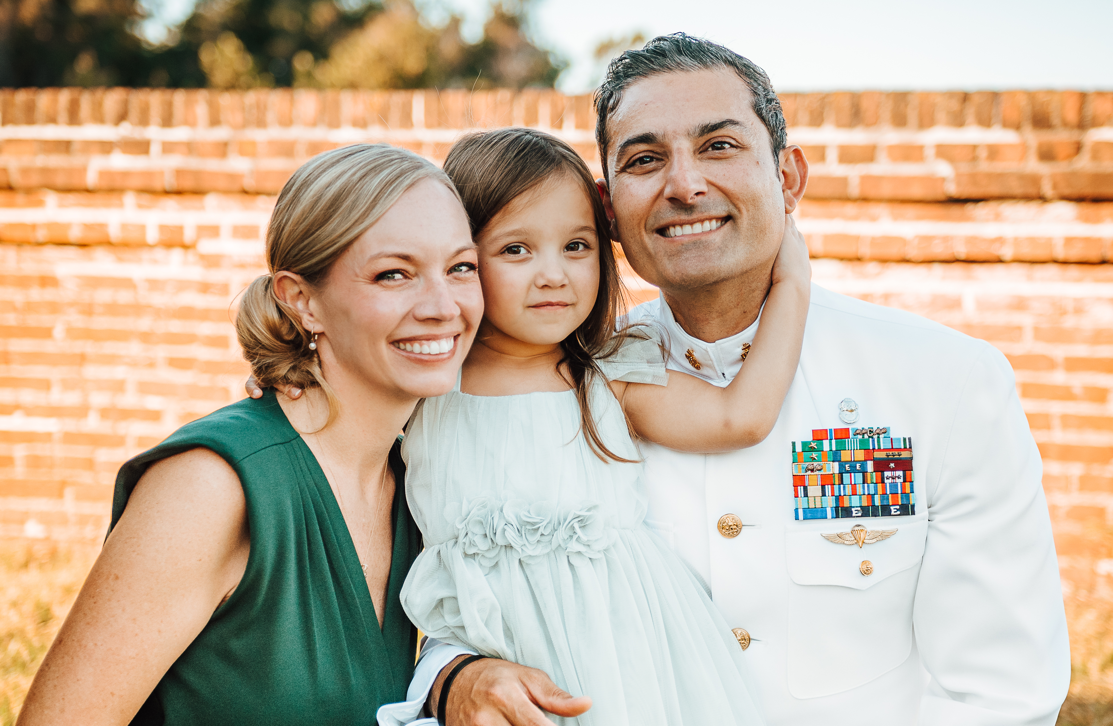 Amy and Chris Perez pose for a family photo with their daughter, Ava - Two adults smile while posing closely with a young child outdoors. The child in a light dress hugs the adult in a white military dress uniform with ribbons. The other adult wears a green dress and leans in beside them. A brick wall fills the background in warm daylight.