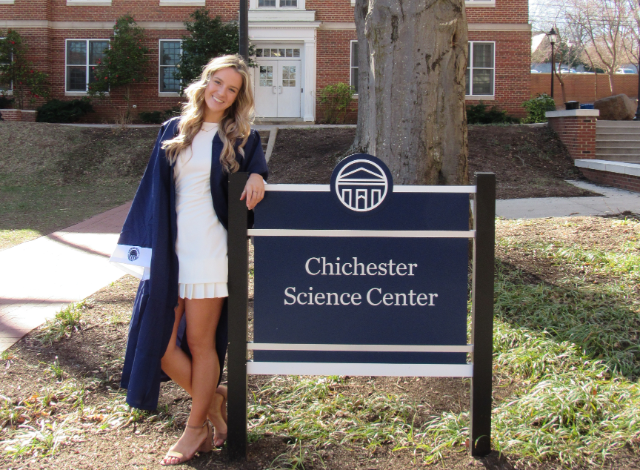Ashley Seiders '25 - A student in a navy graduation gown smiles while standing outdoors. She leans against a sign that reads “Chichester Science Center.” A brick campus building and a large tree are visible in the background. Sunlight falls across the walkway and landscaped planting beds.