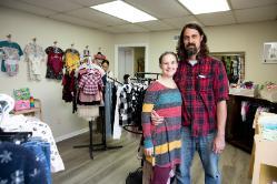 Drs. Adam and Sarai Blincoe pose in their new store, A Giving Tree, located in Farmville, Virginia - A couple stands smiling inside a small children’s clothing shop. Racks of kids’ outfits fill the room, with more clothing displayed on the walls. The man in a red plaid shirt has his arm around the woman in a striped dress. Another shopper browses clothing in the background near a doorway.
