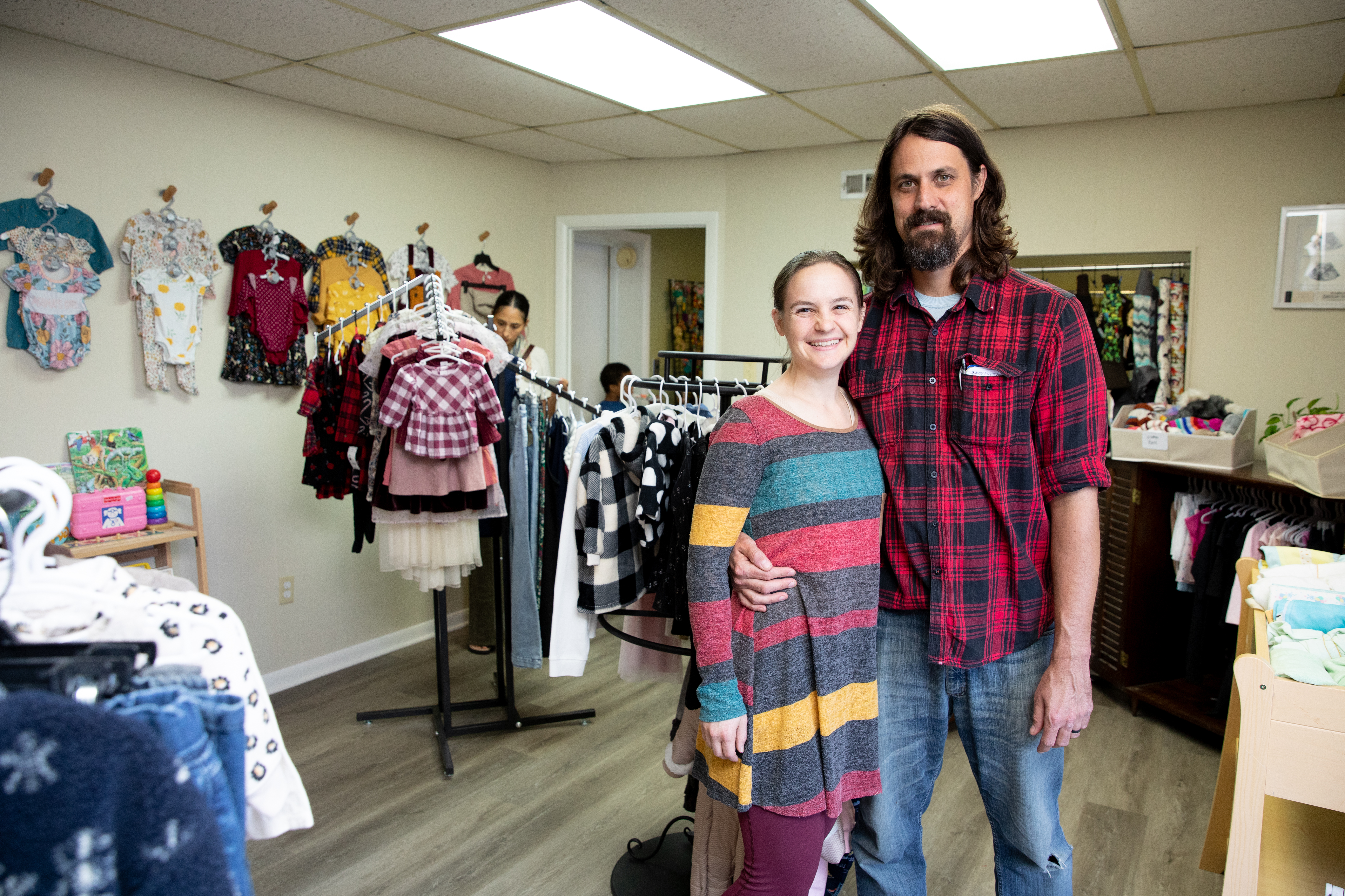 Drs. Adam and Sarai Blincoe pose in their new store, A Giving Tree, located in Farmville, Virginia - A couple stands smiling inside a small children’s clothing shop. Racks of kids’ outfits fill the room, with more clothing displayed on the walls. The man in a red plaid shirt has his arm around the woman in a striped dress. Another shopper browses clothing in the background near a doorway.