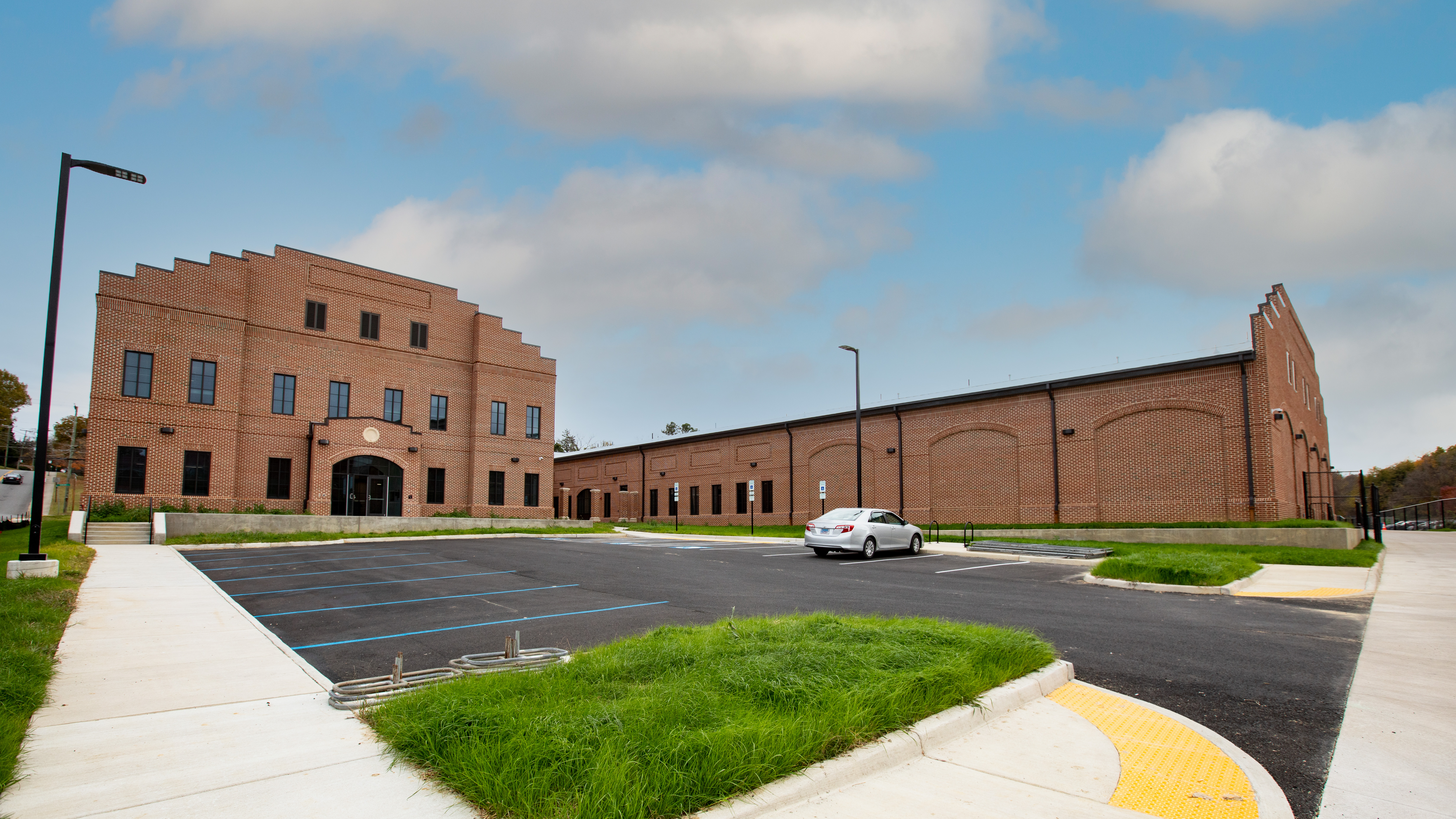 Charter Hall - A wide view of a large brick building with a stepped facade and arched entryway. An empty parking lot stretches across the foreground with a single car parked near the building. Sidewalks, curb cuts, and patches of green grass border the lot. A blue sky with scattered clouds hangs above the building and light poles.