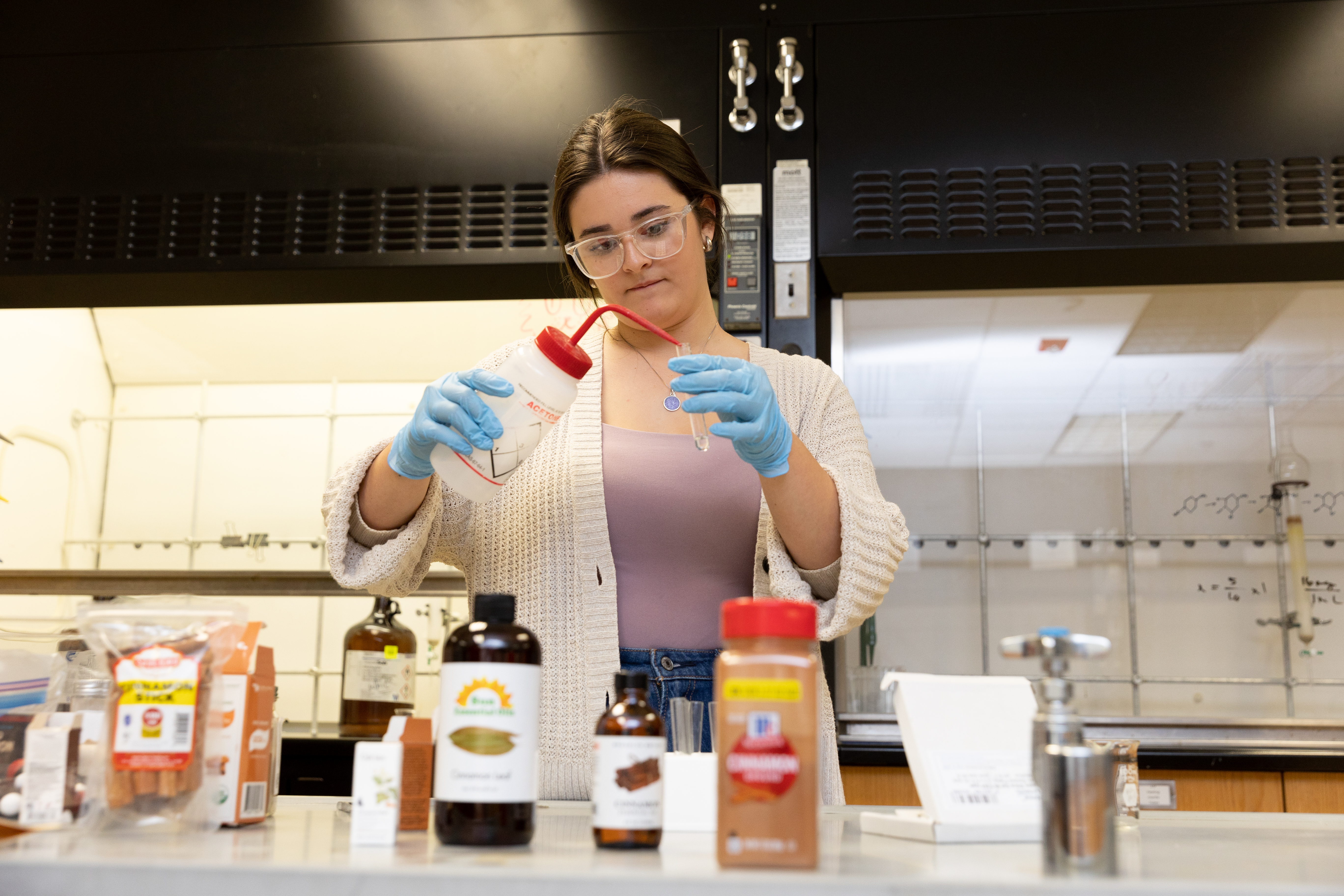Moorea Whitfield '26 testing various cinnamon brand in Longwood's chemistry lab - A student wearing safety goggles and blue gloves works at a lab bench. They pour liquid from a wash bottle into a small test tube. Bottles of cinnamon and other ingredients sit on the counter in the foreground. A fume hood and lab equipment are visible behind them.