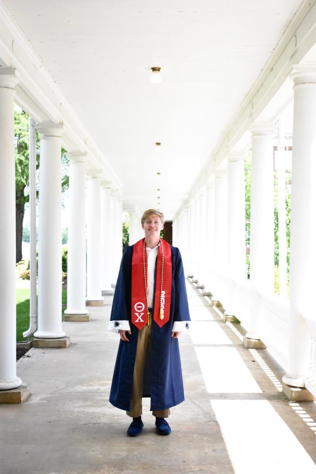 William Poch ’25 - Blue wildcat logos appear on the banner above. A graduate stands centered in a white colonnade on campus. They wear a navy commencement robe with a red stole and cords. Sunlight and long shadows stretch down the walkway between columns. The graduate smiles toward the camera in a full-length portrait.