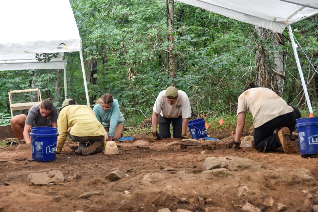 Longwood students at Red Hill work together to excavate the grounds of a cabin that once housed enslaved people at Red Hill Plantation. - Five students kneel and crouch in a dirt excavation area beneath two white canopy tents. They use small tools and blue buckets while carefully clearing soil in marked sections. Rocky ground and orange flag markers outline the dig area in the foreground. Dense gr