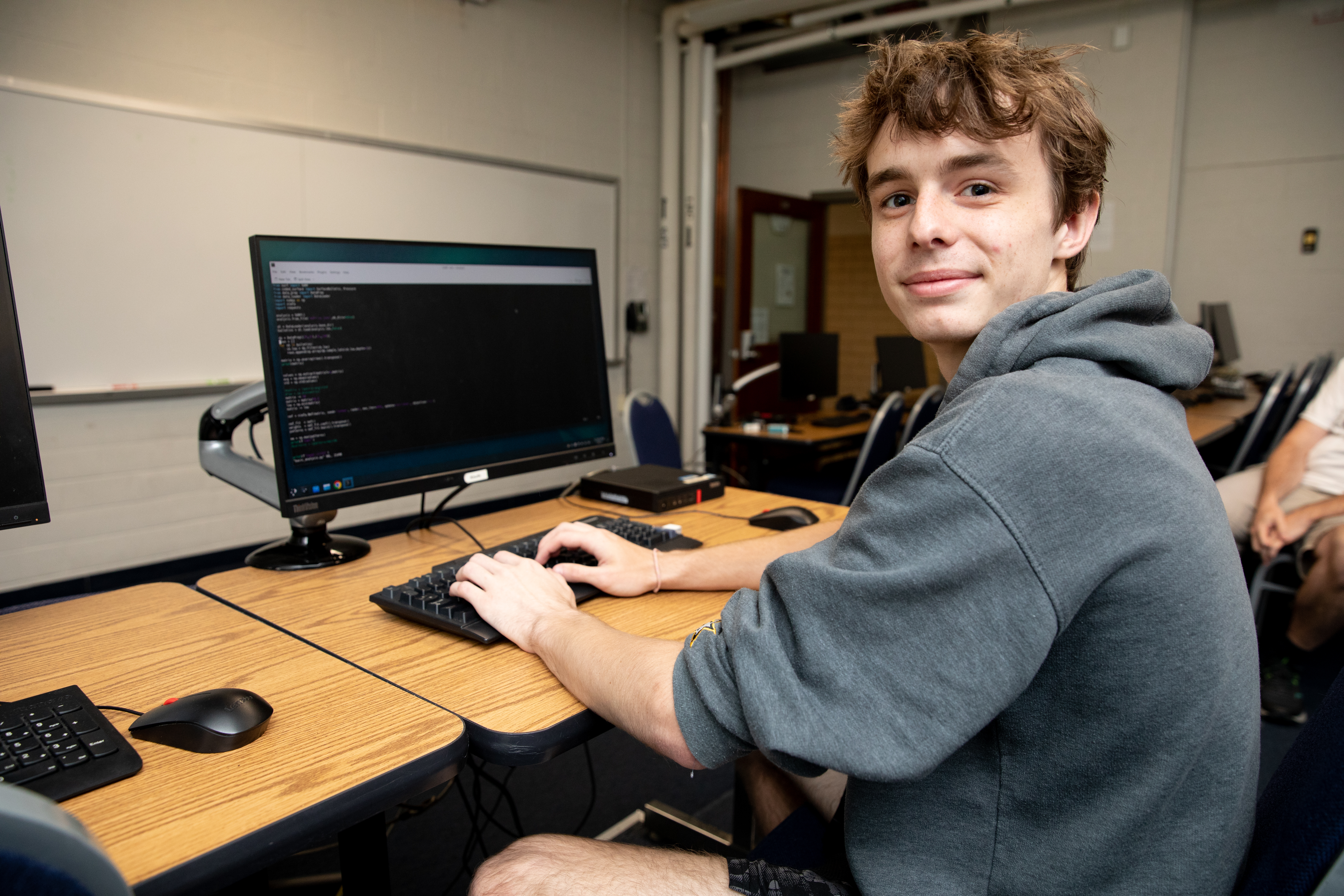 Eli Stark ’26 working at his computer - A student in a gray hoodie sits at a computer workstation in a lab. Code is displayed on the monitor as he types on a keyboard. He turns toward the camera and smiles while seated at the desk. Other computers and classroom equipment are visible in the background.