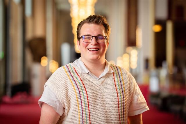 Headshot of Aidan Brown '26 - A smiling person wearing glasses stands in a hallway with warm, out-of-focus lights behind them. They wear a white shirt under a cream knit sweater vest with colorful striped trim. The background shows a red carpeted corridor with columns and soft bokeh lighting. The portrait is centered and brightly lit with a shallow depth of field.