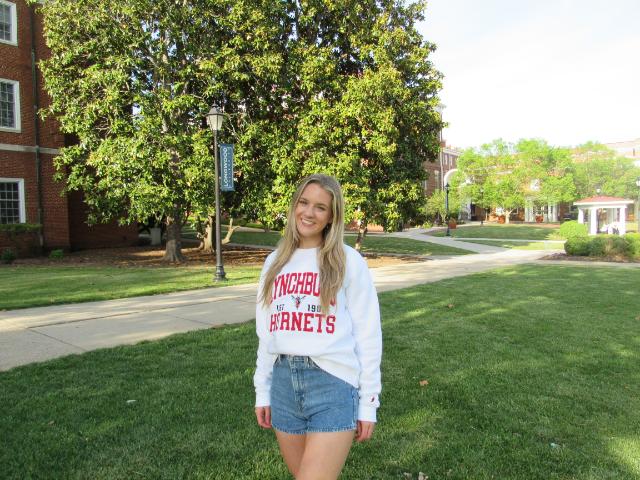 Ashley Seiders ’25 - Student smiles on a campus lawn beside a walkway and large green trees. She wears a white “Lynchburg Hornets” sweatshirt and denim shorts. Brick buildings and a Longwood banner on a lamppost appear in the background. Sunlight brightens the quad with paths leading toward campus buildings.