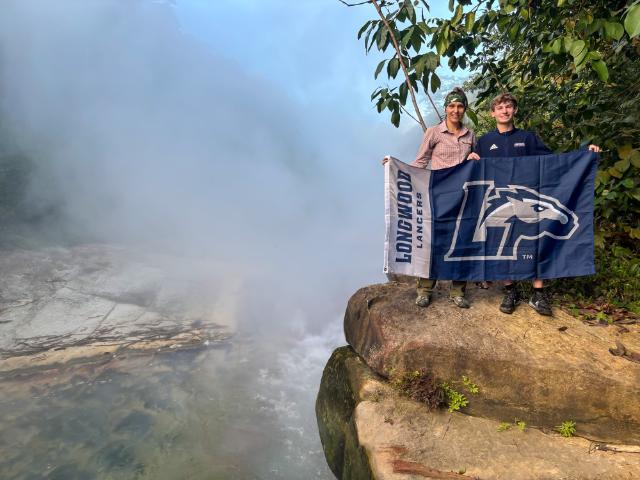Becky Schnekser ’05 and Andrew Schaefer ’26 - write alt text Thought for a couple of seconds  Two people stand on a rocky ledge beside a steaming river in a lush forest. They hold a blue Longwood Lancers flag stretched out in front of them. Thick mist rises from the water, partially obscuring the riverbed and trees. Green foliage and overhanging branches frame the scene on the right.