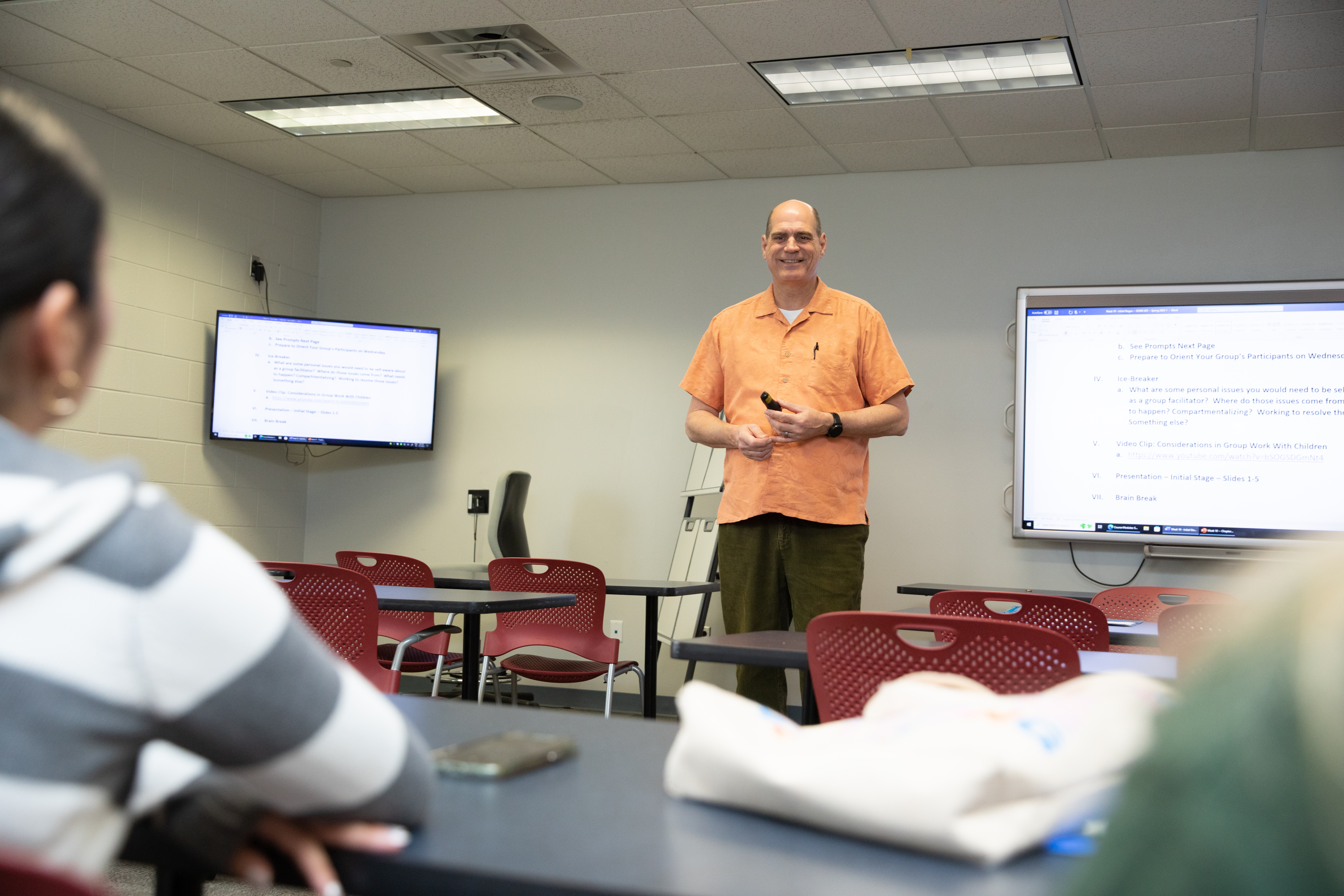 - An instructor stands at the front of a classroom, smiling and holding a presentation remote. Two wall-mounted screens display a document or lesson outline. Students sit at tables in the foreground, slightly out of focus. Red chairs and classroom furniture fill the room under fluorescent ceiling lights.