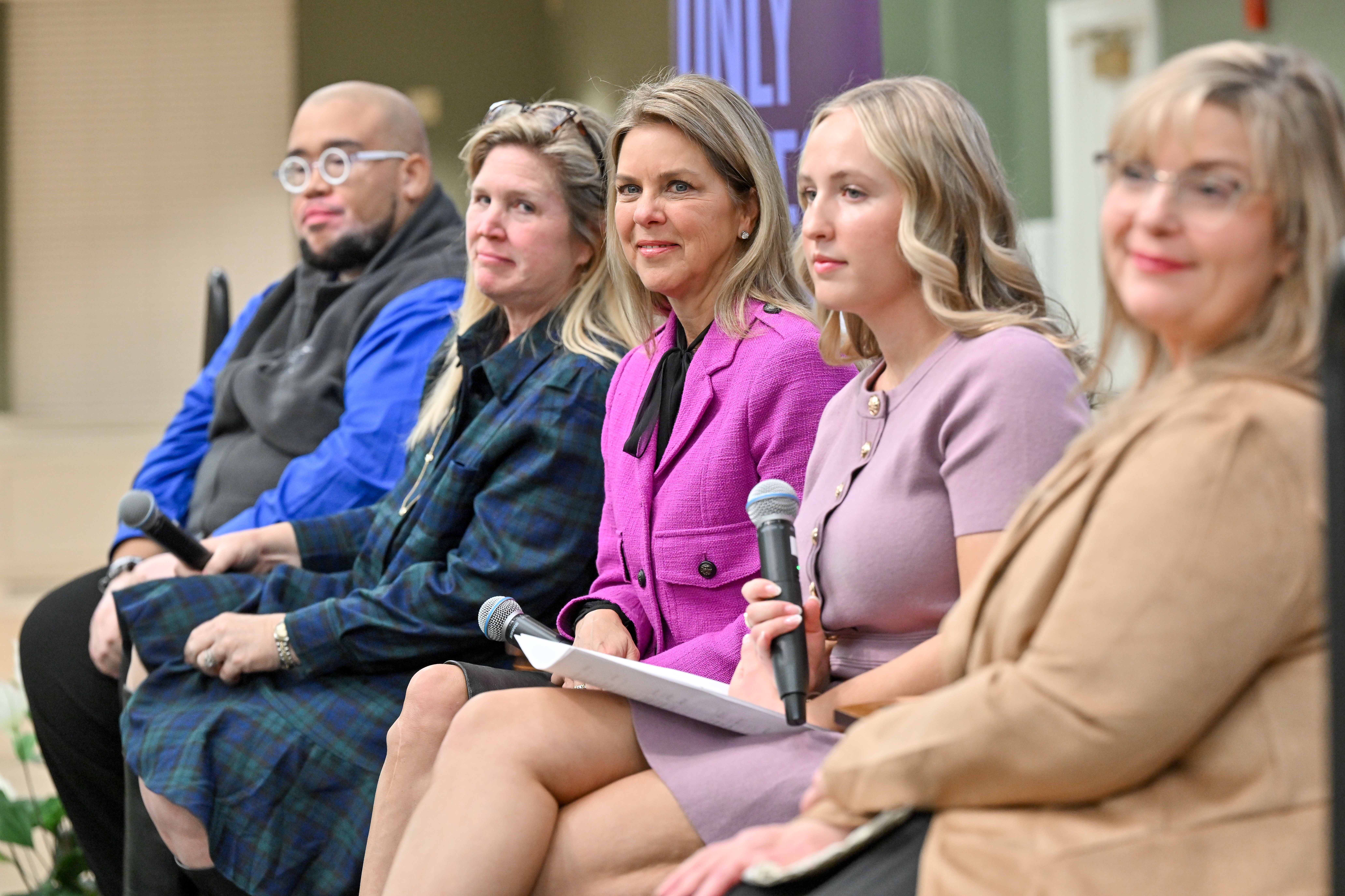 Fentanyl awareness event panelists listen on as an audience member asks a question - Five panelists sit in a row during a discussion, facing an audience off camera. A woman in a lavender dress holds a microphone while listening. A woman in a bright magenta jacket sits beside her, with other panelists holding microphones. The indoor event space is softly blurred in the background.