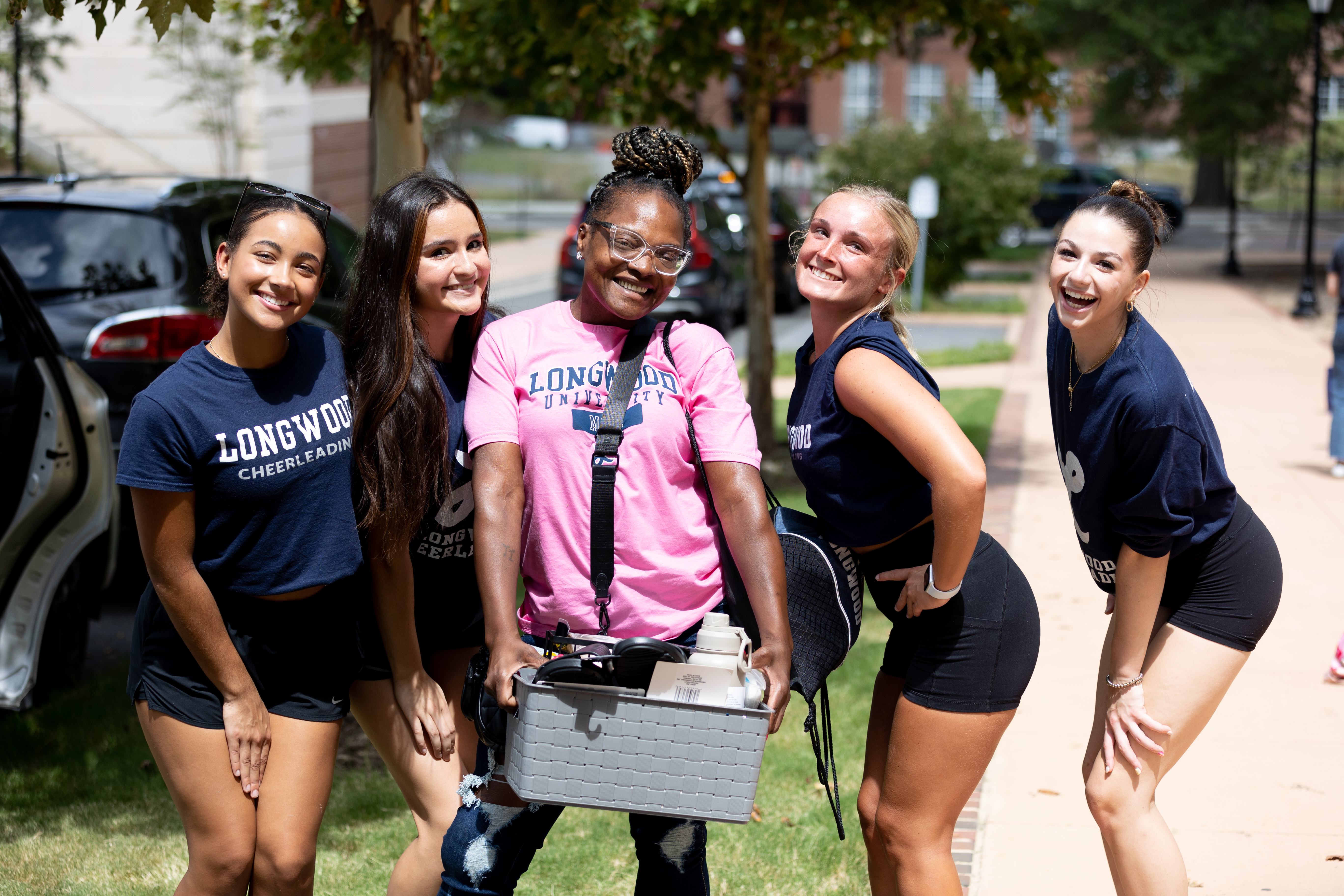 Freshman move-in with volunteers - Five students smile and pose together on a sidewalk during move-in. Two wear Longwood cheerleading shirts and athletic shorts. A student in a pink Longwood University shirt holds a bin of dorm supplies. Cars, trees, and residence hall sidewalks line the background.