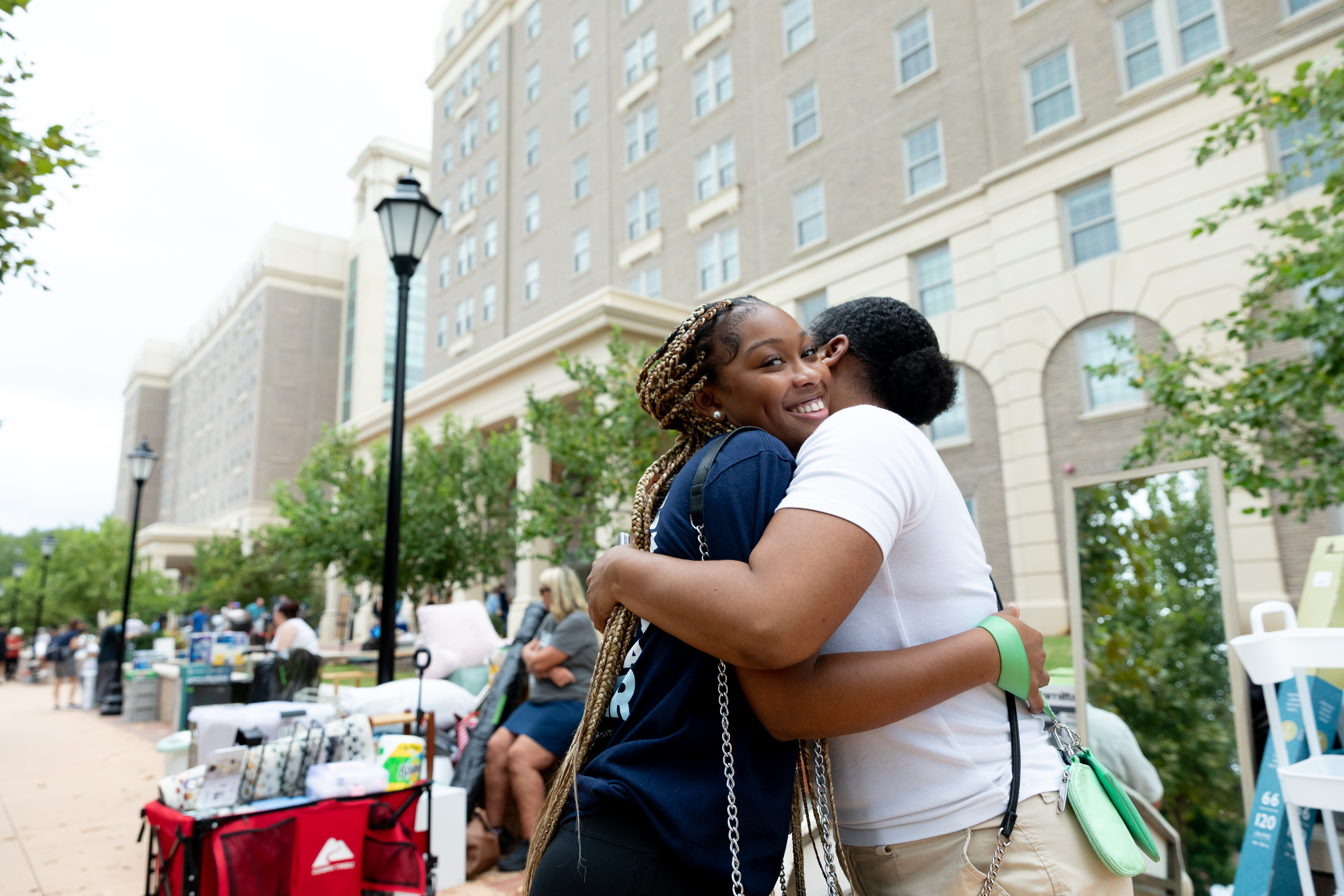 Family member hugs their student in from of Moss Hall - Two students hug outside a Longwood residence hall during move-in. One student smiles toward the camera while being embraced. Move-in carts and boxes line the sidewalk behind them. The tall residence hall building and trees frame the scene.