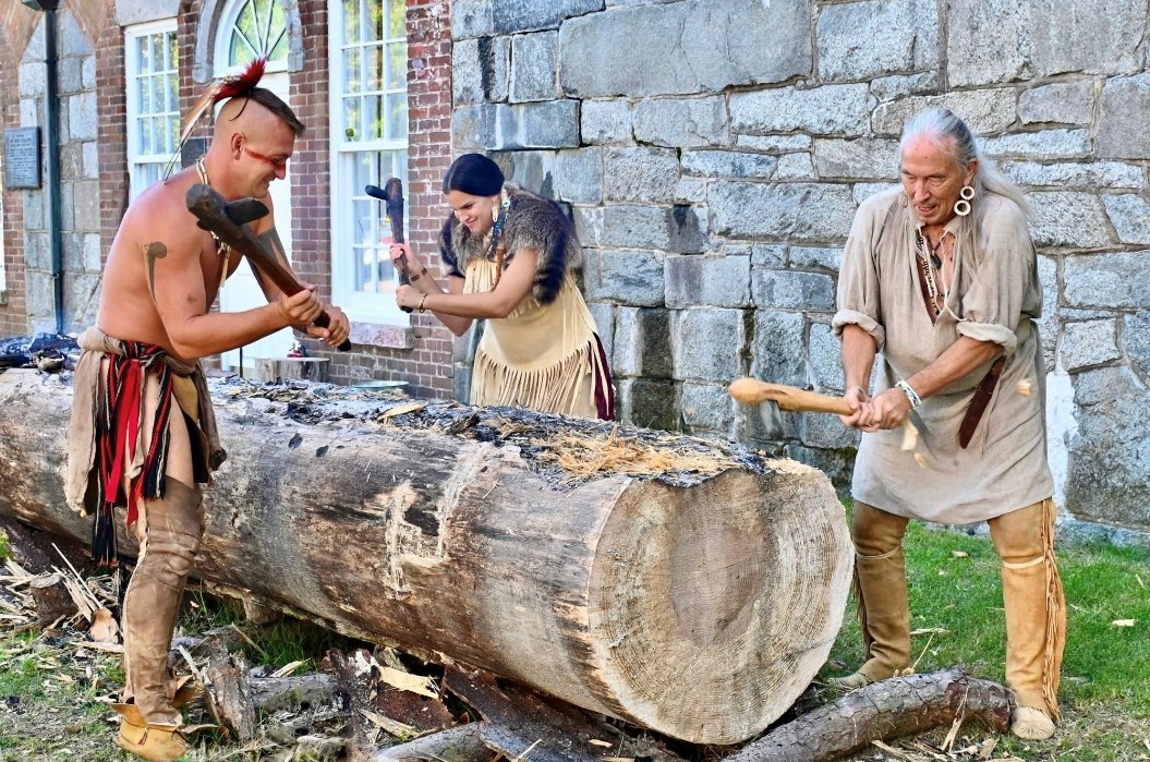 Russell Reed (on left) cutting into a log with swings of his axe, recreating the Native American art form of constructing canoes. - Three reenactors in period clothing carve a large log with axes. One person steadies the log while two others swing axes in mid-strike. Wood chips and shavings scatter across the log and the grass. A stone-and-brick building wall with windows stands behind them.