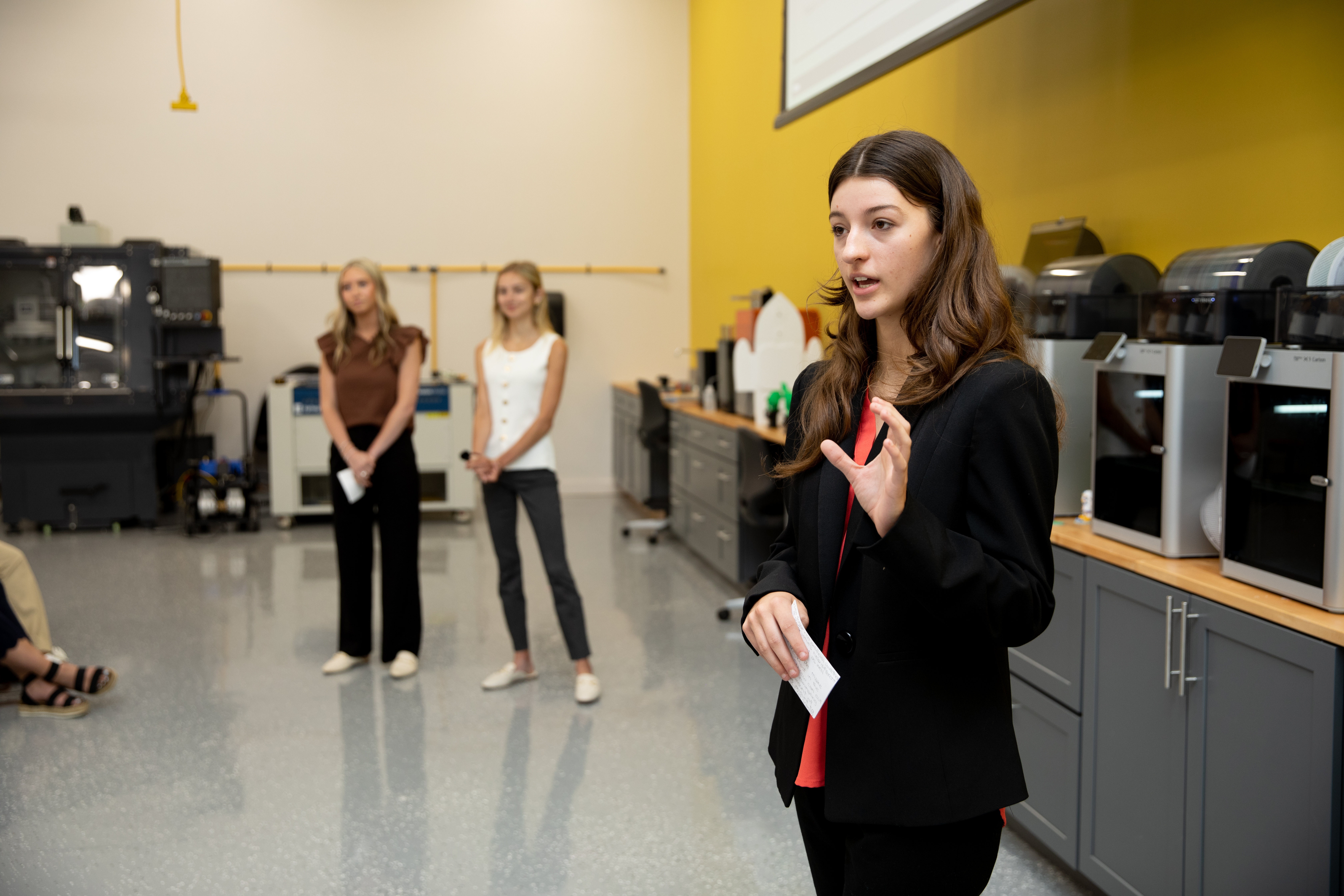 The FitBrew team won third place and $200 for their product: a Keurig-compatible coffee pod spiked with 20 grams of protein that gives a time-pressed person coffee and breakfast in one cup. - Student presents in a technology lab, gesturing while speaking. 3D printers line a counter along the wall behind her. Two students stand in the background near industrial equipment. A projector screen and yel