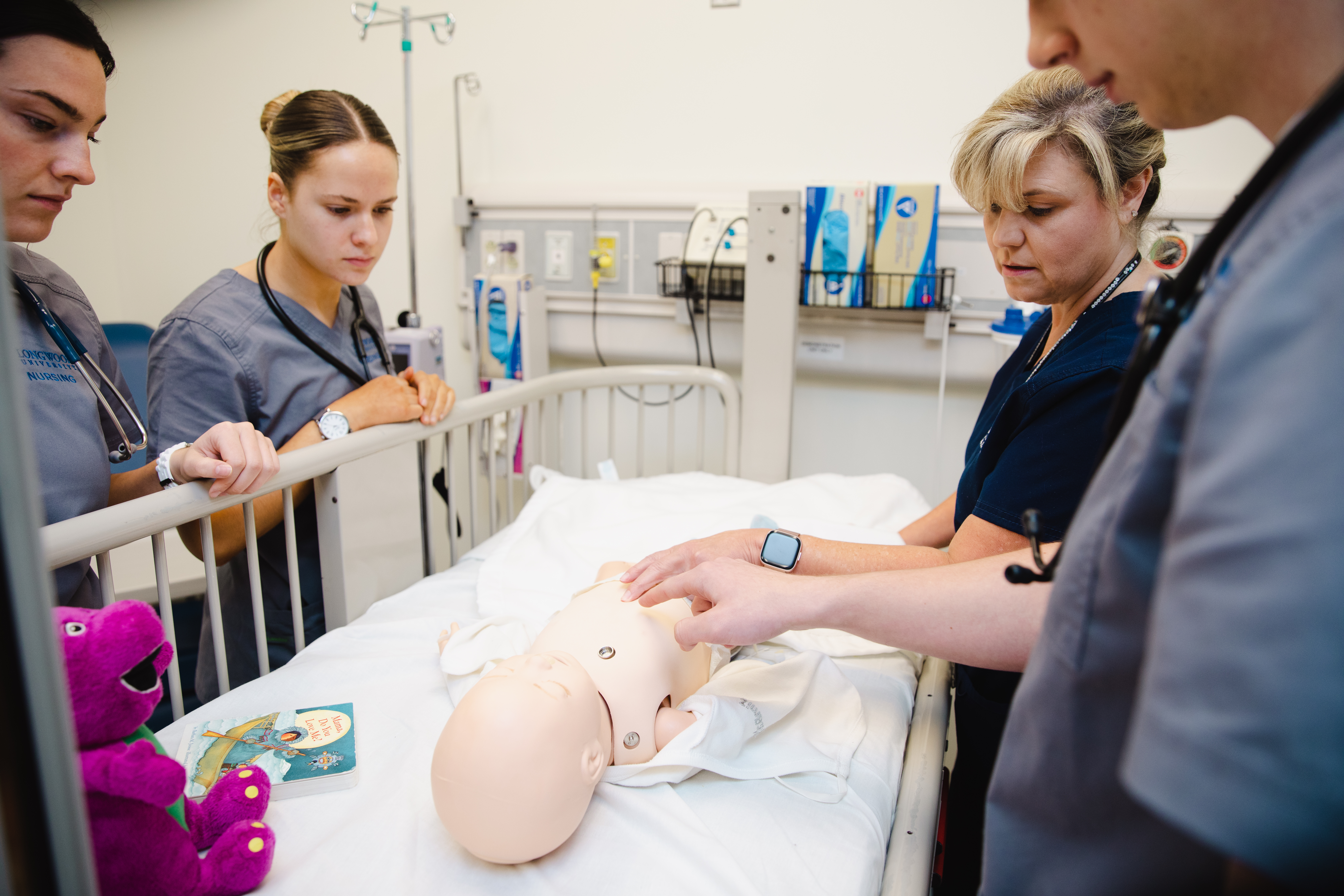 - A nursing instructor and students stand around a hospital bed in a simulation lab. The instructor demonstrates care on a baby manikin lying under a white sheet. Students in scrubs with stethoscopes watch closely from the bedside rail. A plush purple dinosaur toy and a children’s book rest near the manikin.