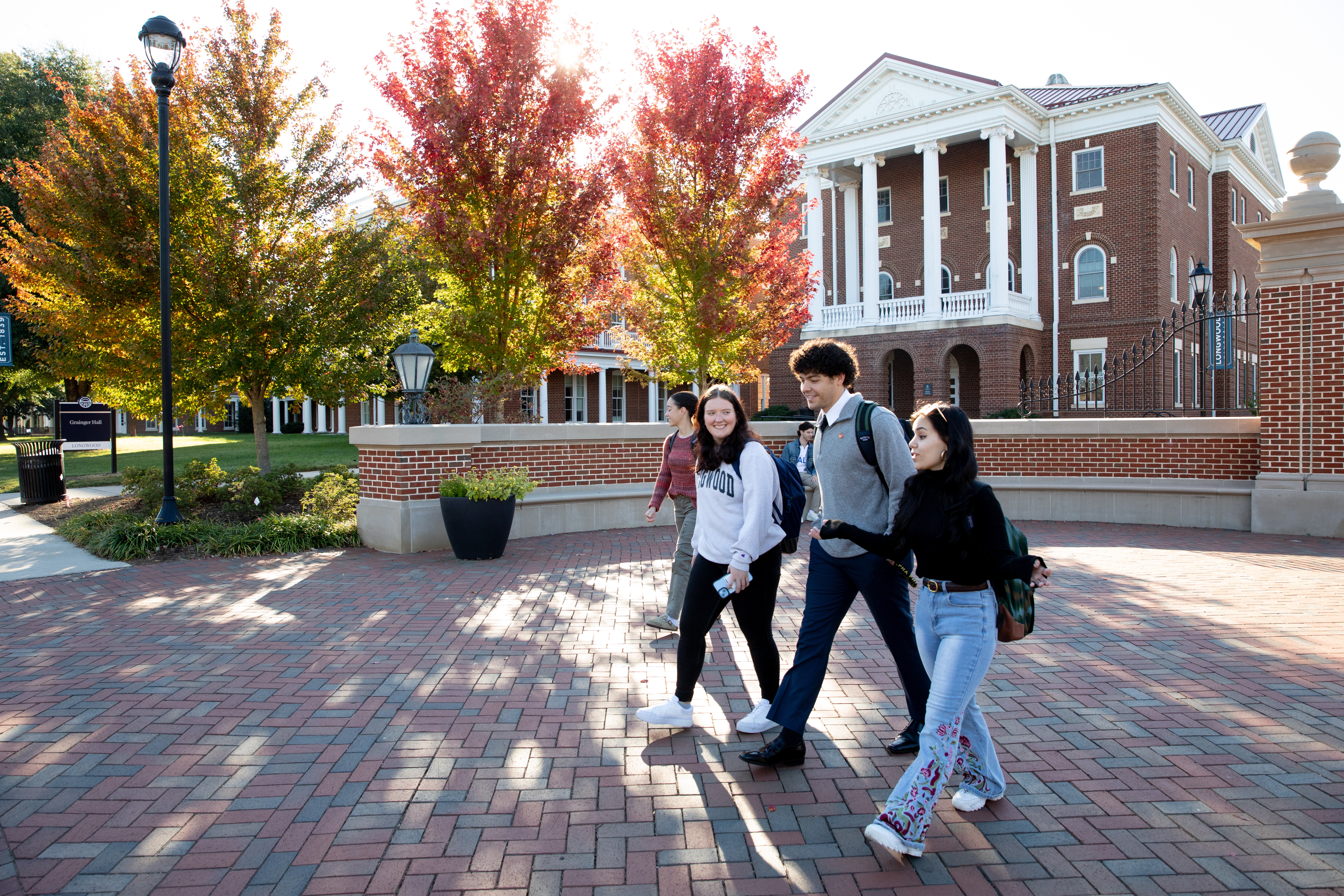 Students walking in front of the Longwood gate in fall - Four students walk and talk across a brick plaza on campus. They wear casual clothes and backpacks as they cross in front of a red-brick building. White columns, an iron fence, and a lamppost frame the scene. Trees with red and green fall leaves glow in the sunlight behind them.