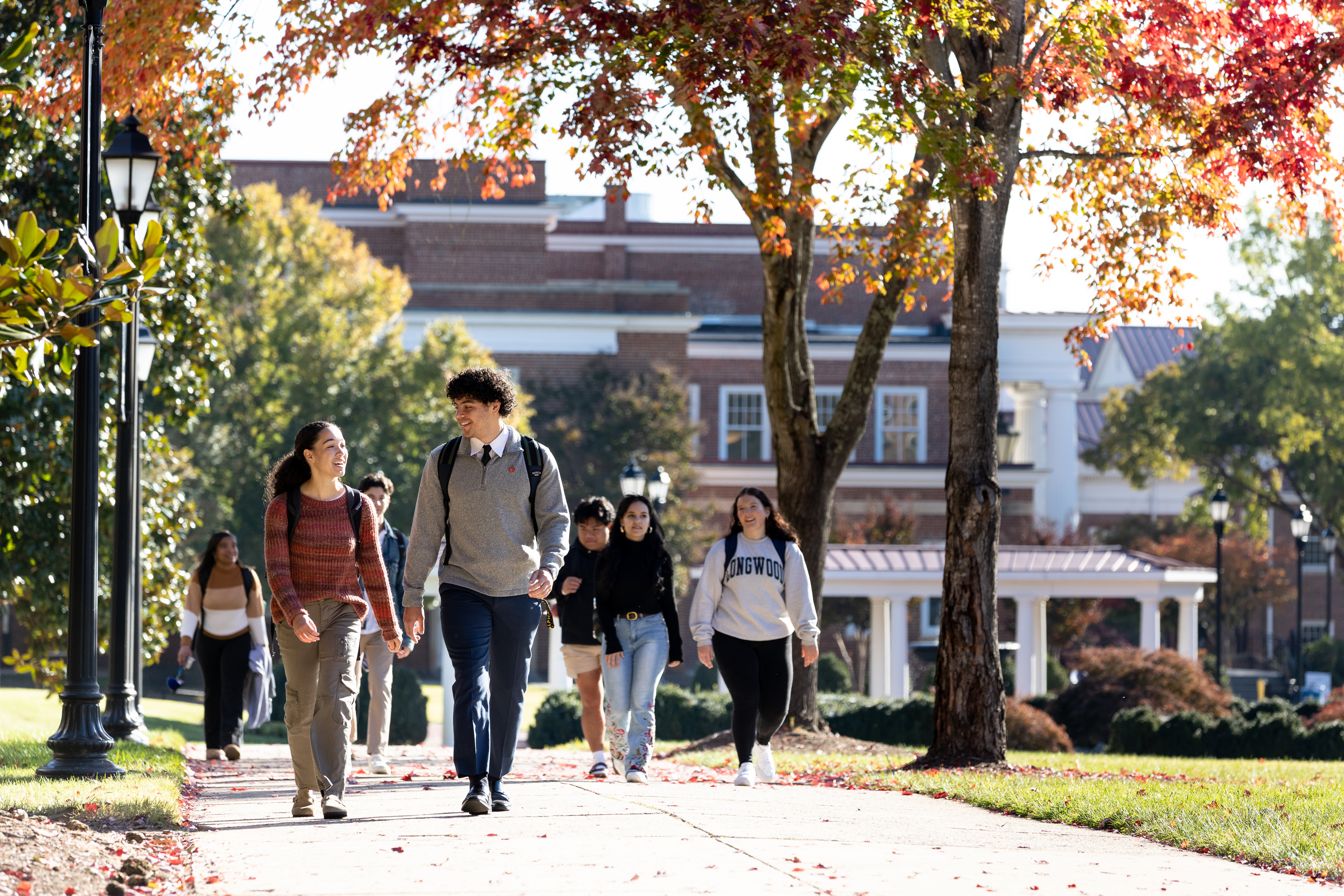 Students walking behind Eason during the fall - Students walk along a campus sidewalk beneath trees with red and orange leaves. Two students with backpacks chat as they lead a small group down the path. A brick academic building and white colonnade appear in the background. Sunlight highlights fallen leaves scattered across the walkway.