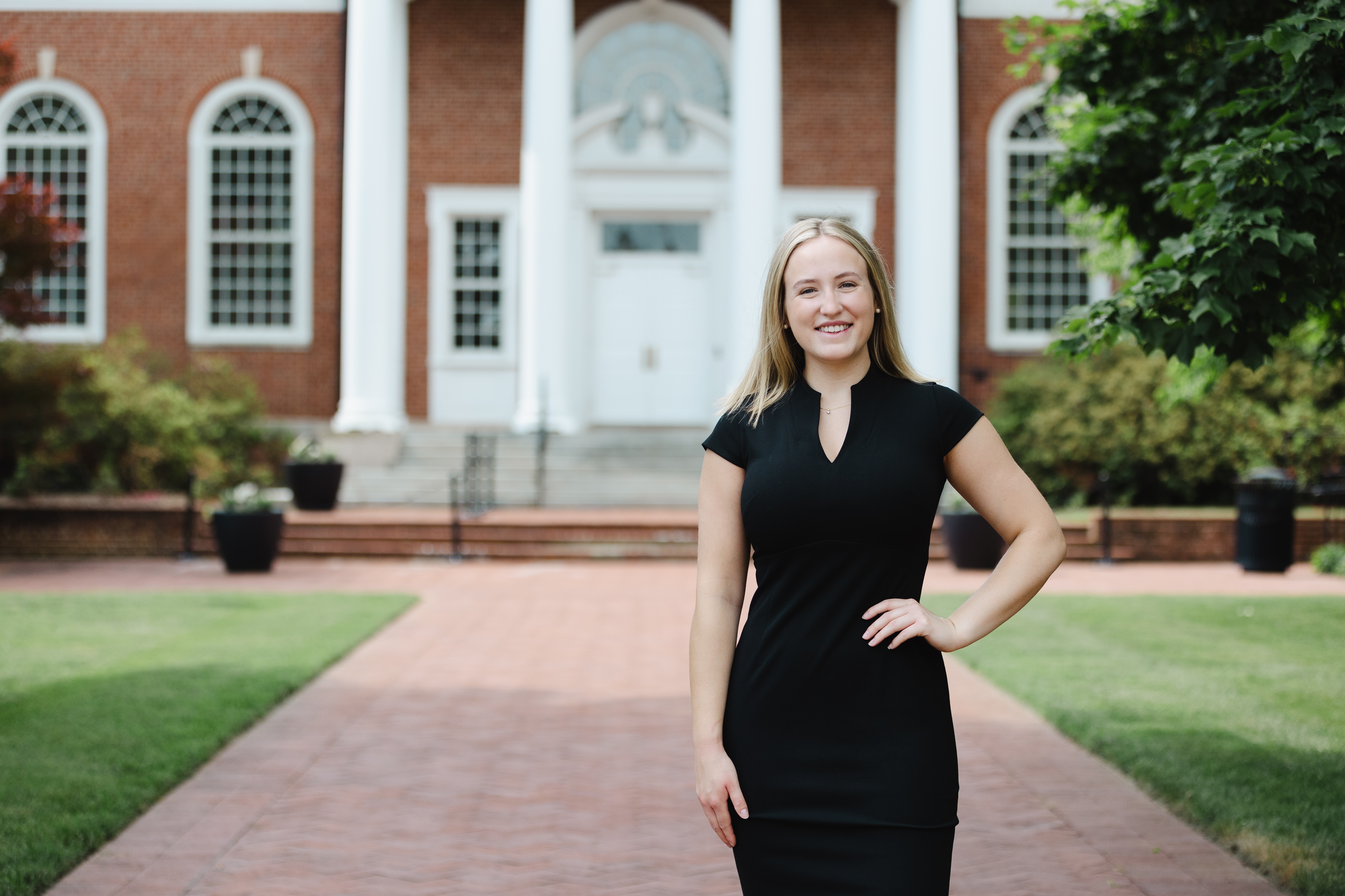Taylor Blount ‘26 - A student stands smiling on a brick walkway in front of a red-brick building. They wear a black dress and pose with one hand on their hip. White columns and arched windows frame the entrance behind them. Green lawns, shrubs, and trees border the walkway.