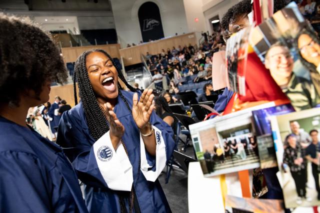 Student grins, laughs, and claps at the sight of their decorated cap. - A student in a navy graduation gown claps and cheers in an auditorium. Other students in gowns stand nearby during a crowded ceremony. A display of photos and notes fills the foreground at right. Rows of seated attendees and the stage area appear in the background.